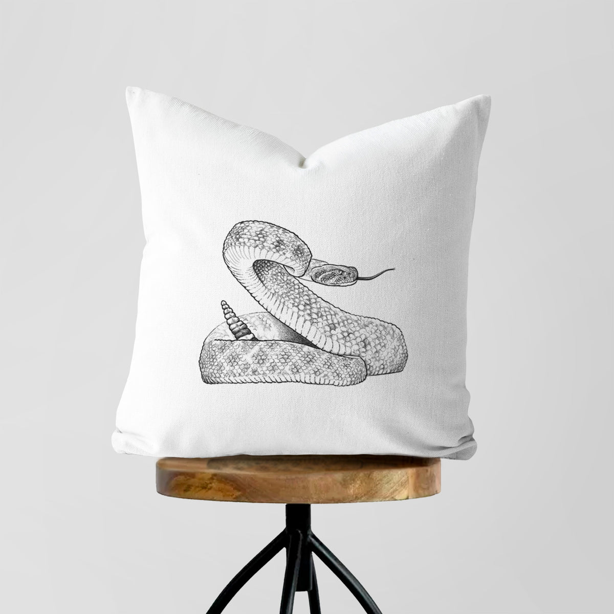 White pillow cover printed with a detailed black ink illustration of a coiled diamondback rattlesnake, on a wooden stool.