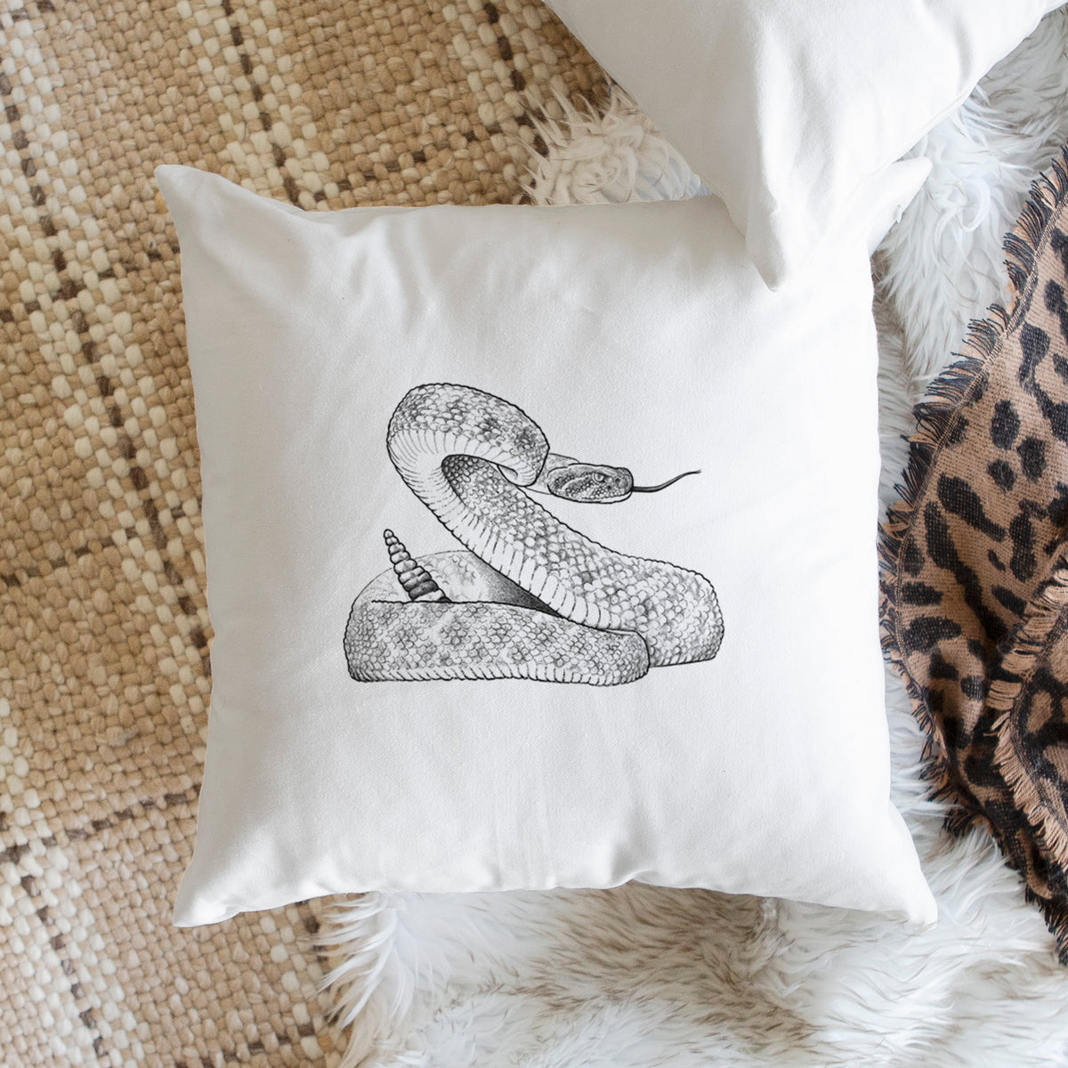 White pillow cover printed with a detailed black ink illustration of a coiled diamondback rattlesnake, on a woven chair.