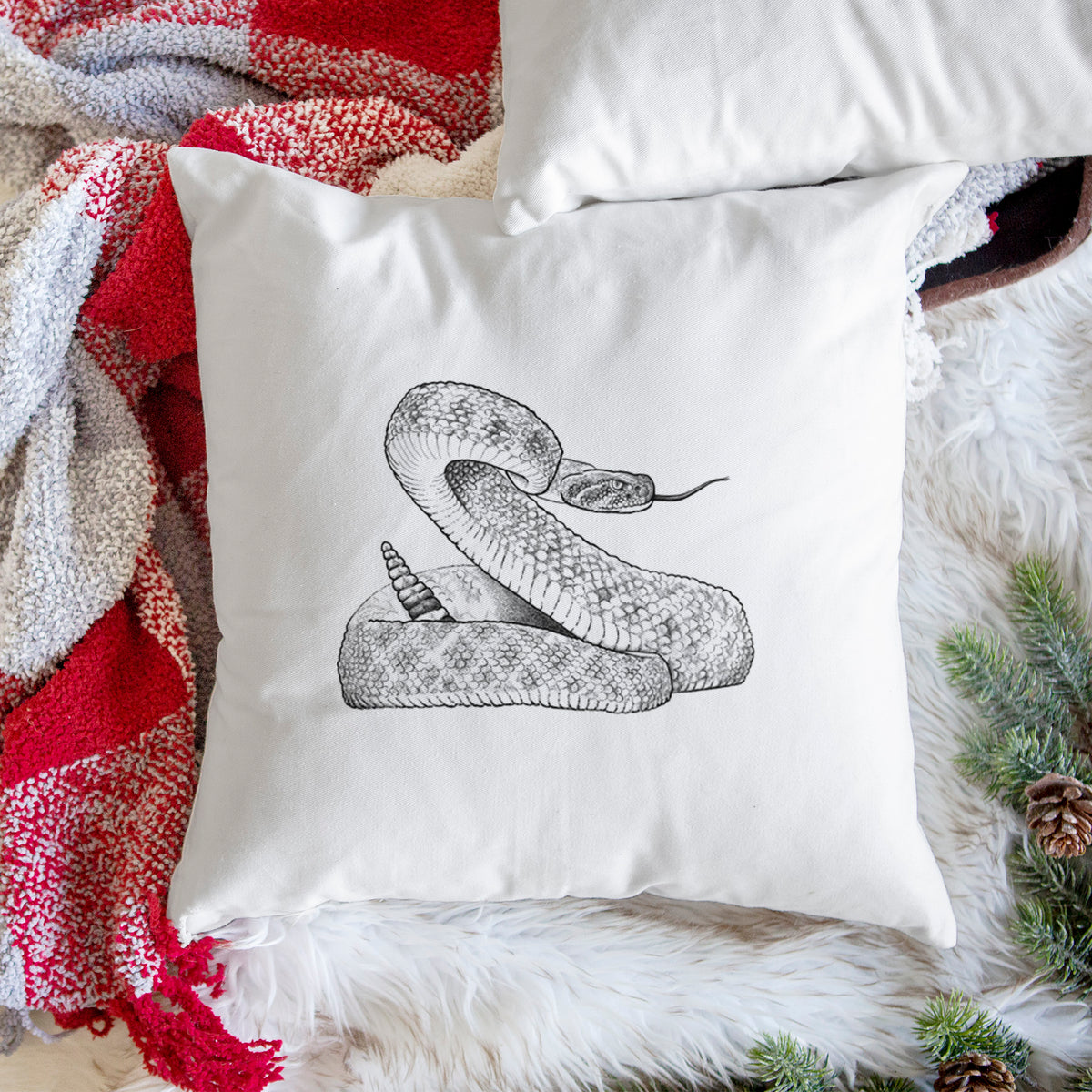 White pillow cover printed with a detailed black ink illustration of a coiled diamondback rattlesnake, on a woven chair.