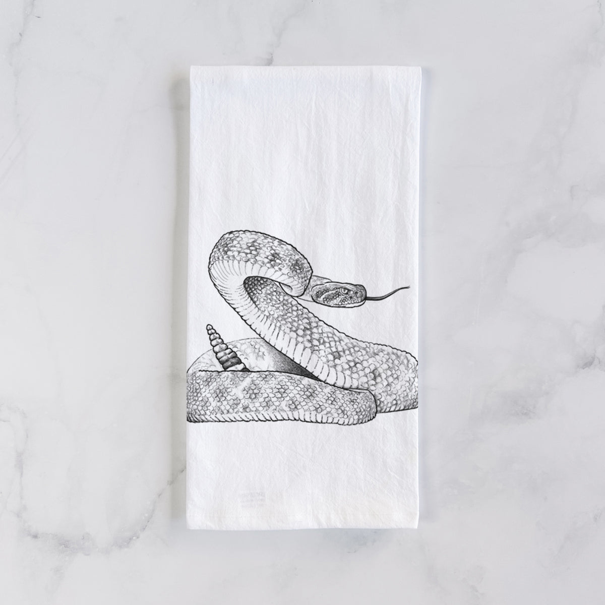 White cotton tea towel laid flat on marble, printed with a detailed black ink illustration of a coiled diamondback rattlesnake.