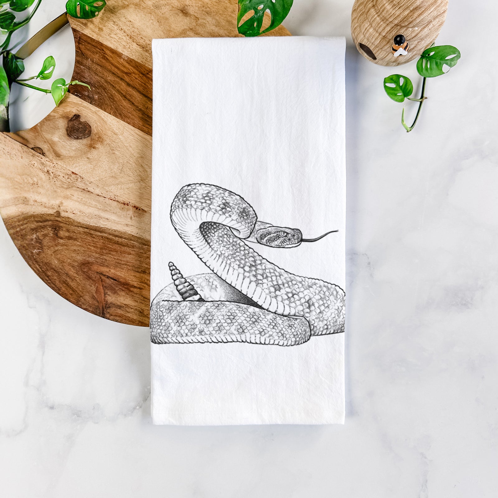 White cotton tea towel printed with a detailed black ink illustration of a coiled diamondback rattlesnake, on a marble counter beside a round wooden cutting board and greenery.
