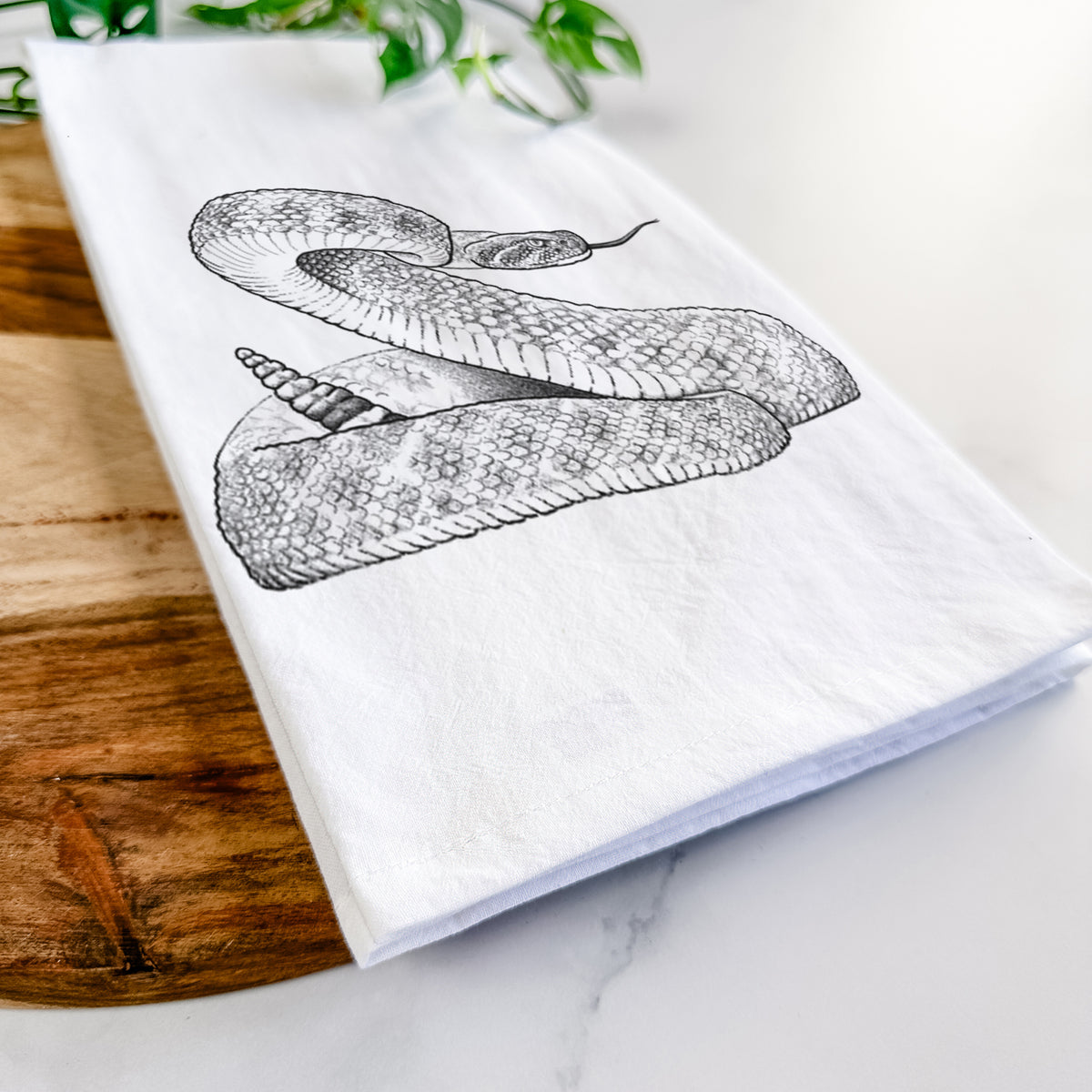 Close-up of a folded white tea towel on a wooden board, showing a detailed black ink illustration of a coiled diamondback rattlesnake.