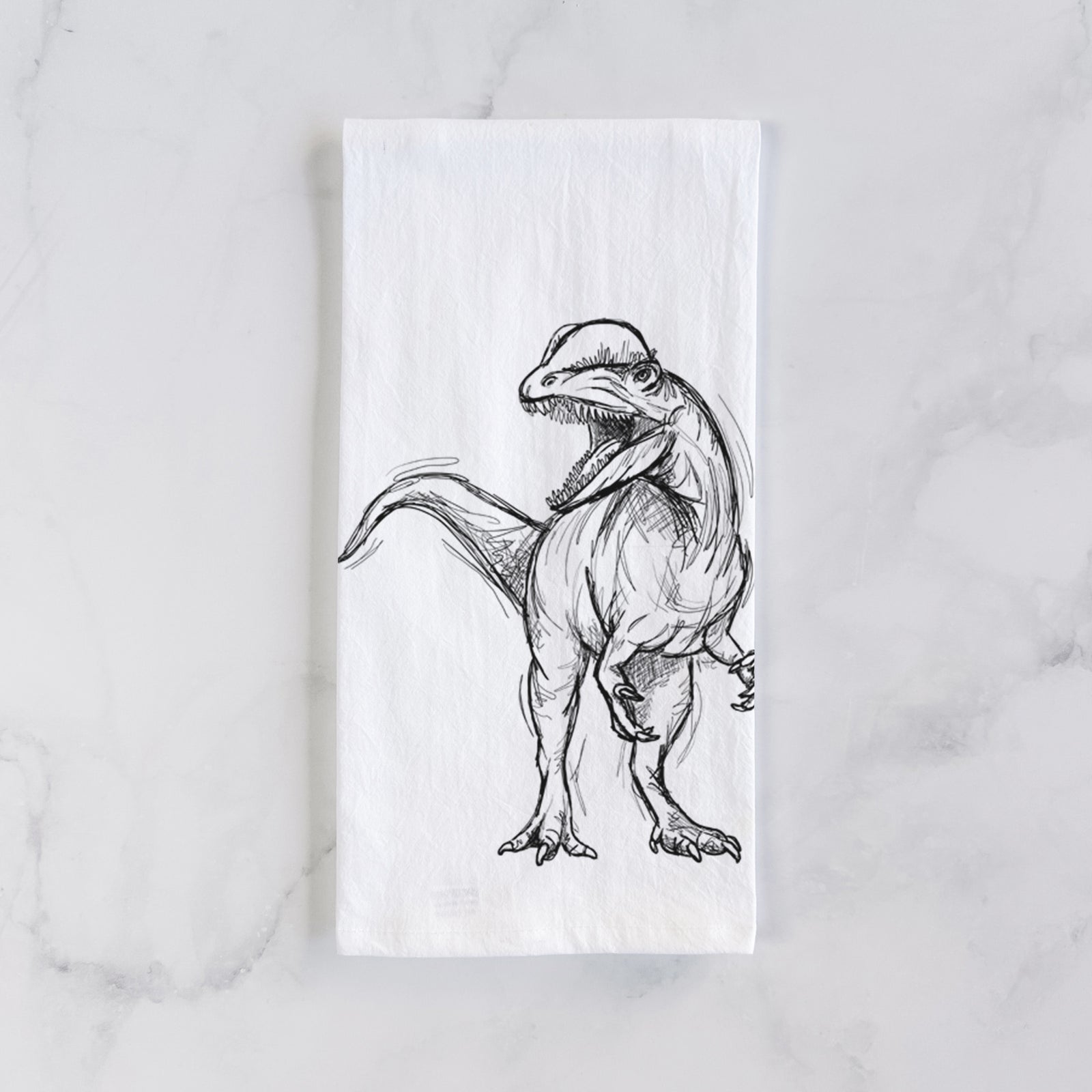 White cotton tea towel printed with a black ink illustration of a Dilophosaurus with its neck frill, walking, on a marble counter beside a round wooden cutting board and greenery.