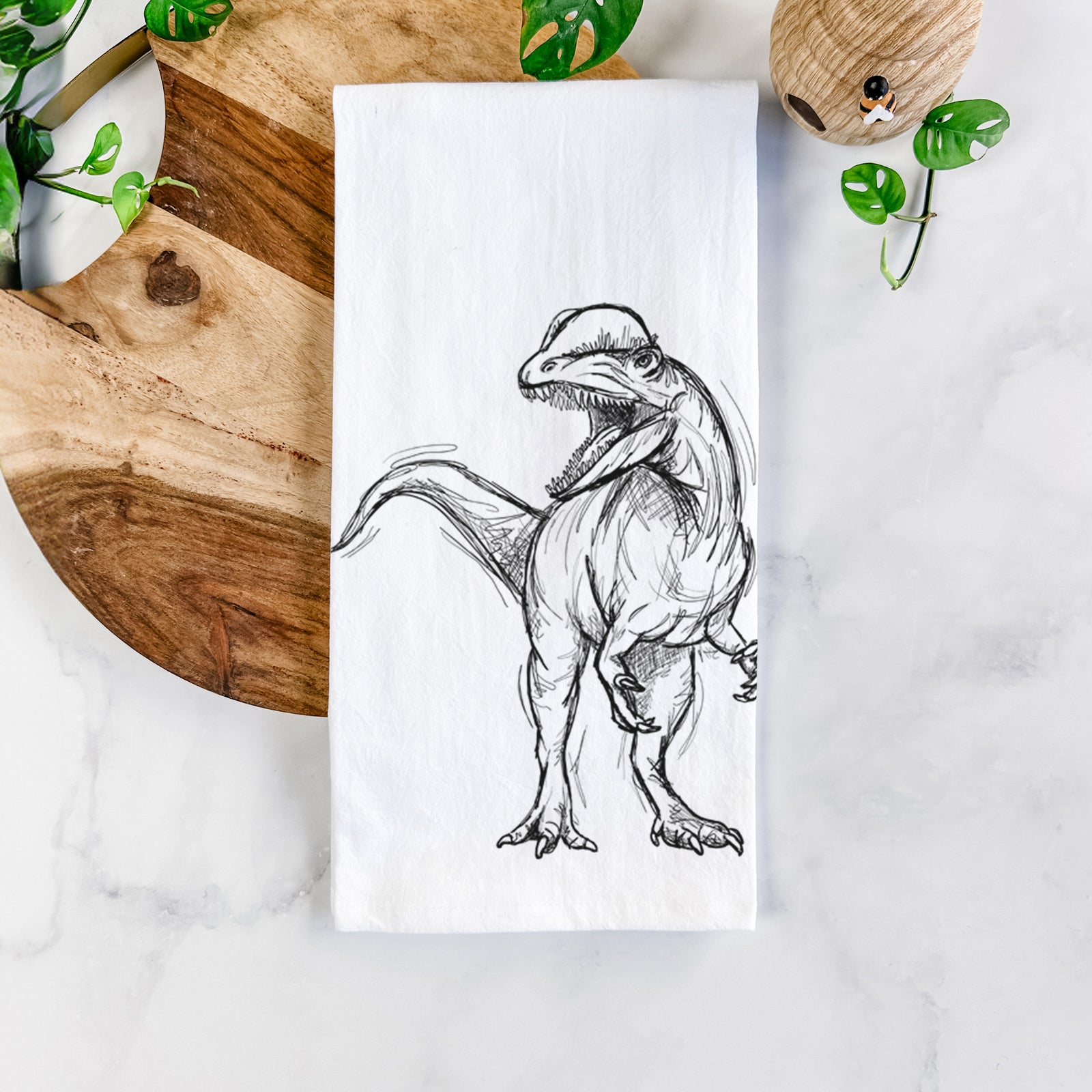 White cotton tea towel printed with a black ink illustration of a Dilophosaurus with its neck frill, walking, on a marble counter beside a round wooden cutting board and greenery.