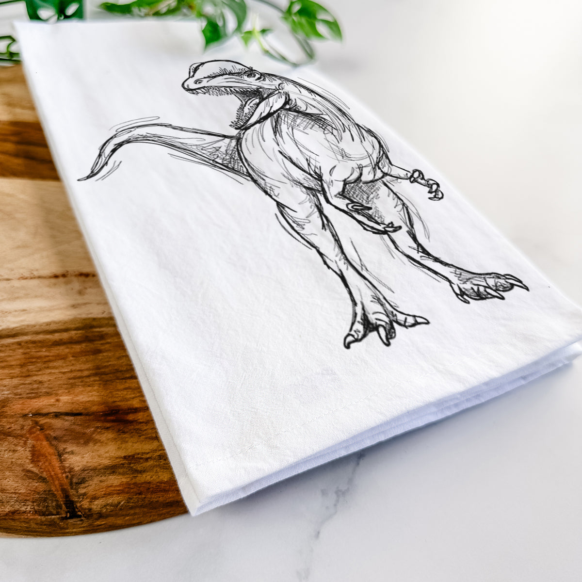 Close-up of a folded white tea towel on a wooden board, showing a black ink illustration of a Dilophosaurus with its neck frill, walking.