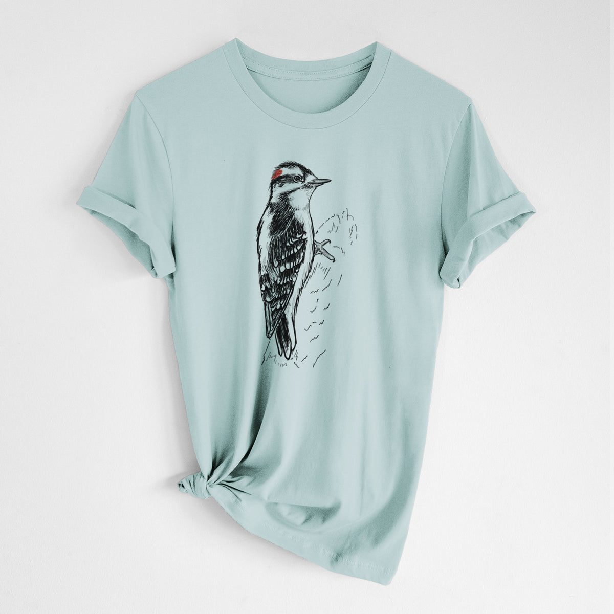 Dusty blue lightweight cotton t-shirt laid flat with the sleeves rolled, printed with a detailed black ink illustration of a downy woodpecker clinging to a tree trunk.