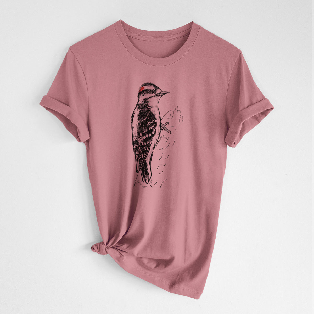 Mauve pink lightweight cotton t-shirt laid flat with the sleeves rolled, printed with a detailed black ink illustration of a downy woodpecker clinging to a tree trunk.