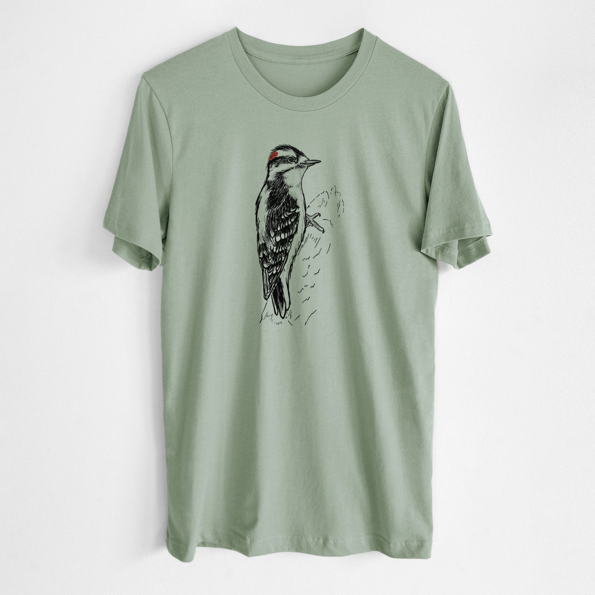 Sage green lightweight cotton t-shirt laid flat, printed with a detailed black ink illustration of a downy woodpecker clinging to a tree trunk.