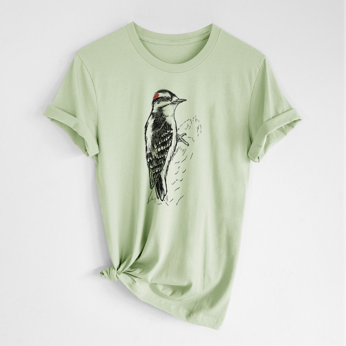 Spring green lightweight cotton t-shirt laid flat with the sleeves rolled, printed with a detailed black ink illustration of a downy woodpecker clinging to a tree trunk.