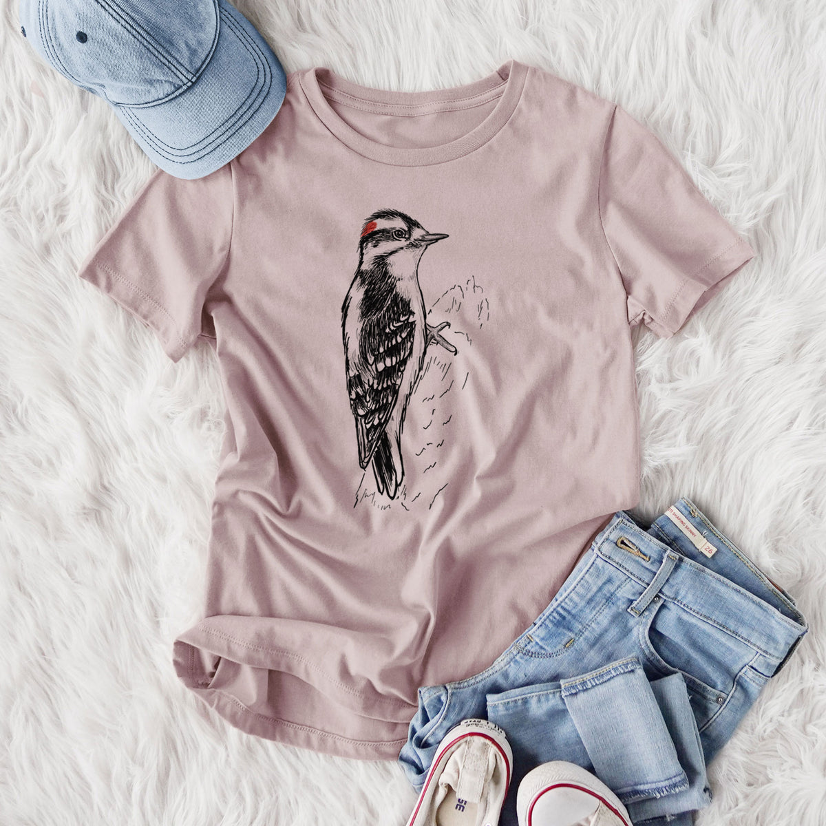 Women's relaxed fit cotton t-shirt printed with a detailed black ink illustration of a downy woodpecker clinging to a tree trunk, styled flat on a white faux fur rug with jeans.
