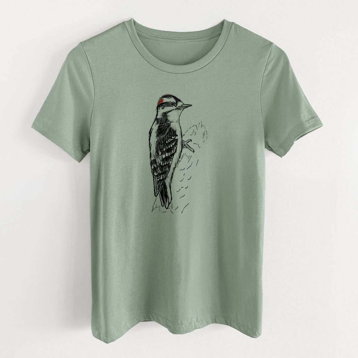 Sage green women's relaxed fit cotton t-shirt laid flat, printed with a detailed black ink illustration of a downy woodpecker clinging to a tree trunk.