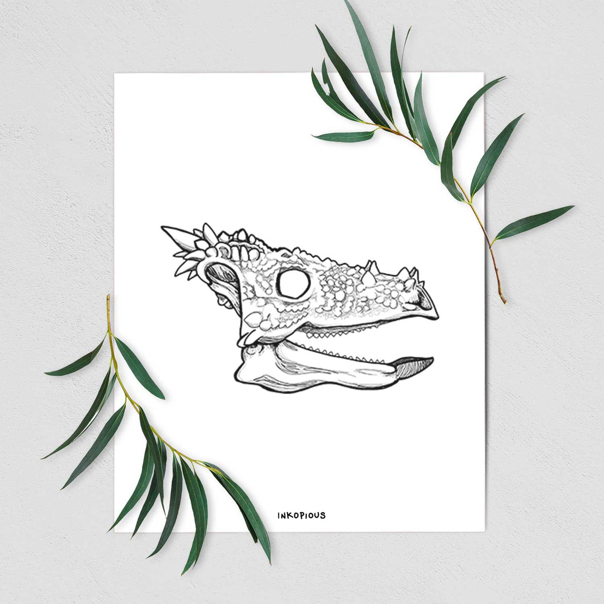 Art print of a black ink illustration of a spiky Dracorex dinosaur skull, shown with eucalyptus leaves.