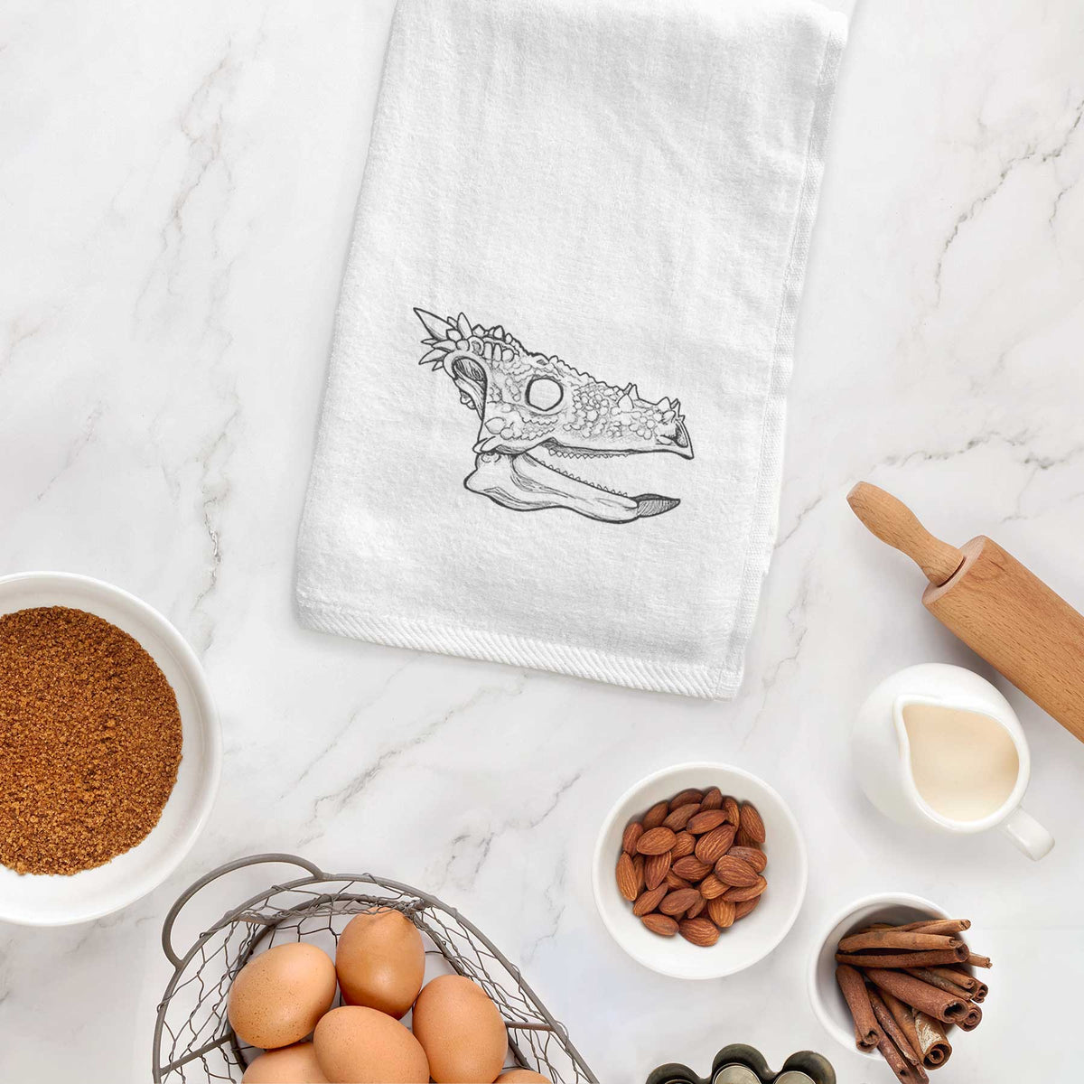White hand towel printed with a black ink illustration of a spiky Dracorex dinosaur skull, hanging on a cabinet door.