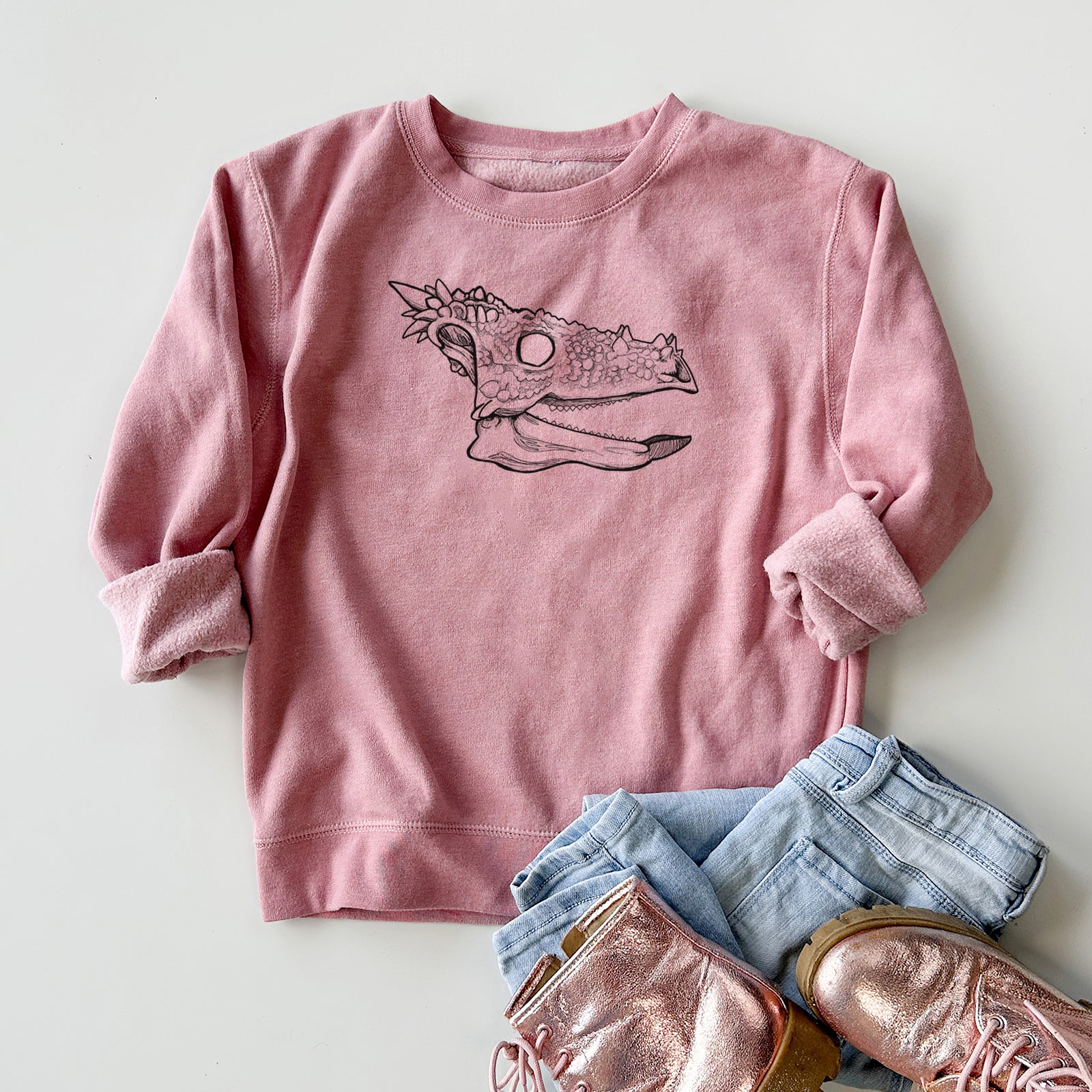 Boy wearing a youth crewneck sweatshirt printed with a black ink illustration of a spiky Dracorex dinosaur skull, outdoors.