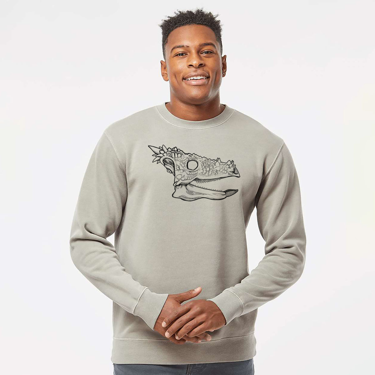 Model wearing a cement grey pigment dyed crewneck sweatshirt printed with a black ink illustration of a spiky Dracorex dinosaur skull.