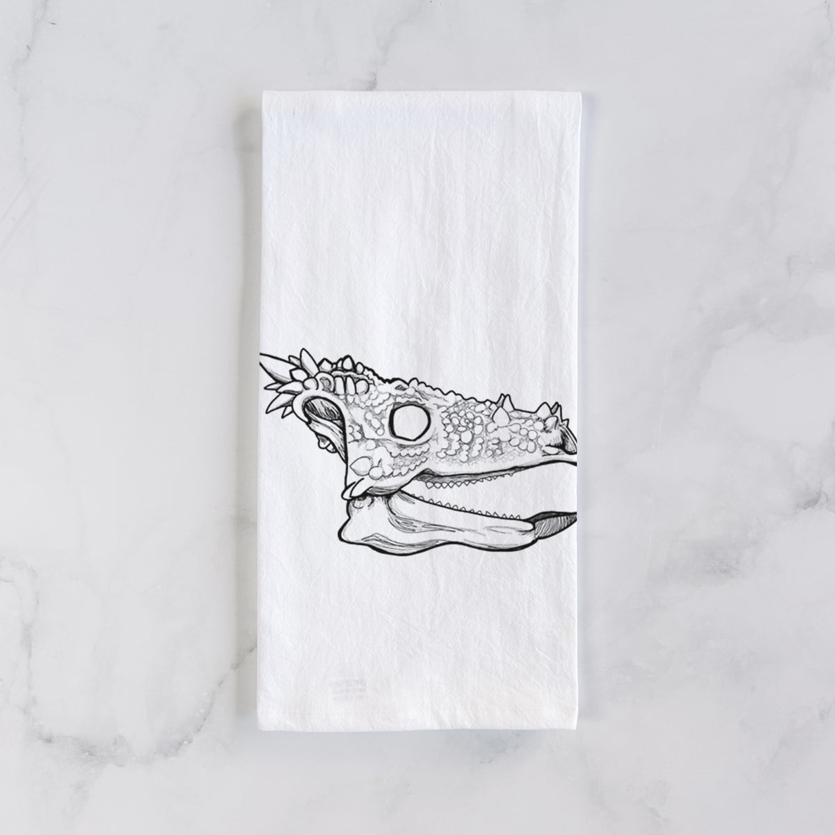 White cotton tea towel laid flat on marble, printed with a black ink illustration of a spiky Dracorex dinosaur skull.