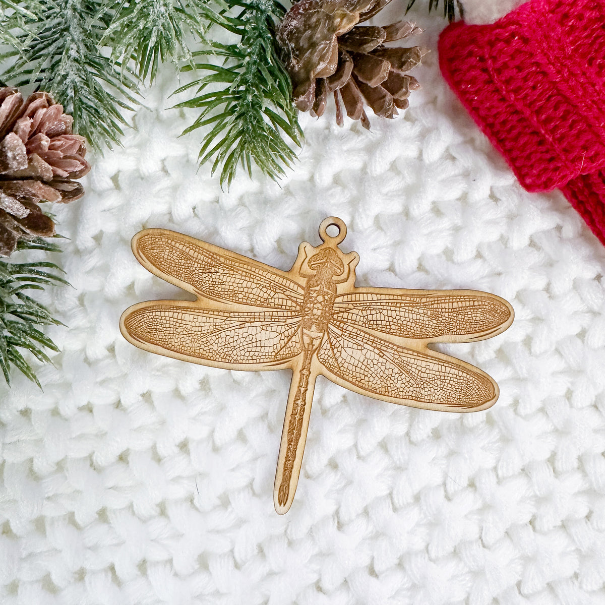 Wooden ornament featuring a detailed engraving of a dragonfly with patterned wings, on a knit blanket with pine cones.