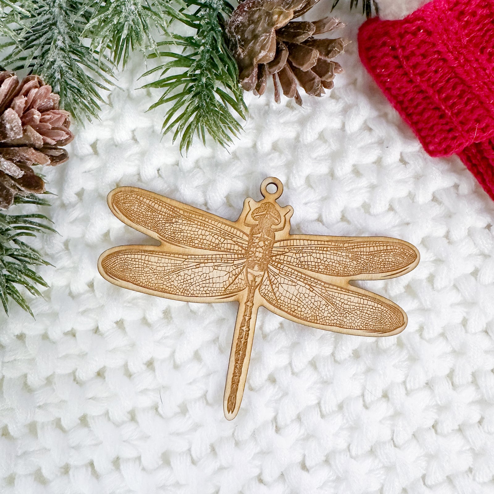 Wooden ornament featuring a detailed engraving of a dragonfly with patterned wings, on a knit blanket with pine cones.