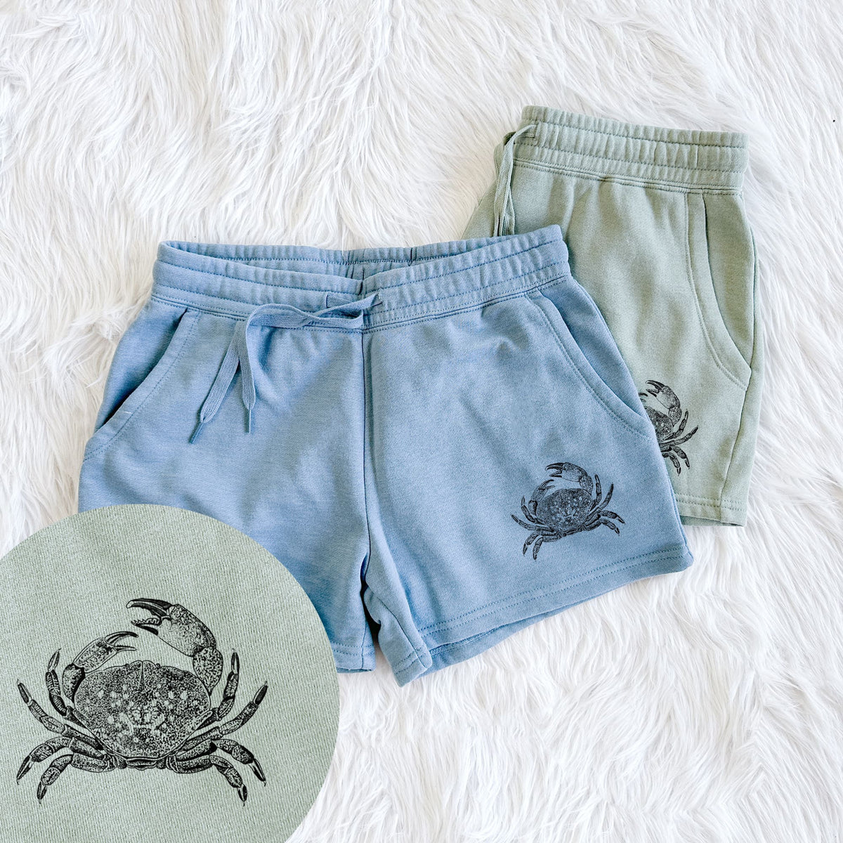 Two pairs of Cali Wave shorts in different colors printed with a detailed black ink illustration of a Dungeness crab with claws raised, seen from above, with a close-up of the print.