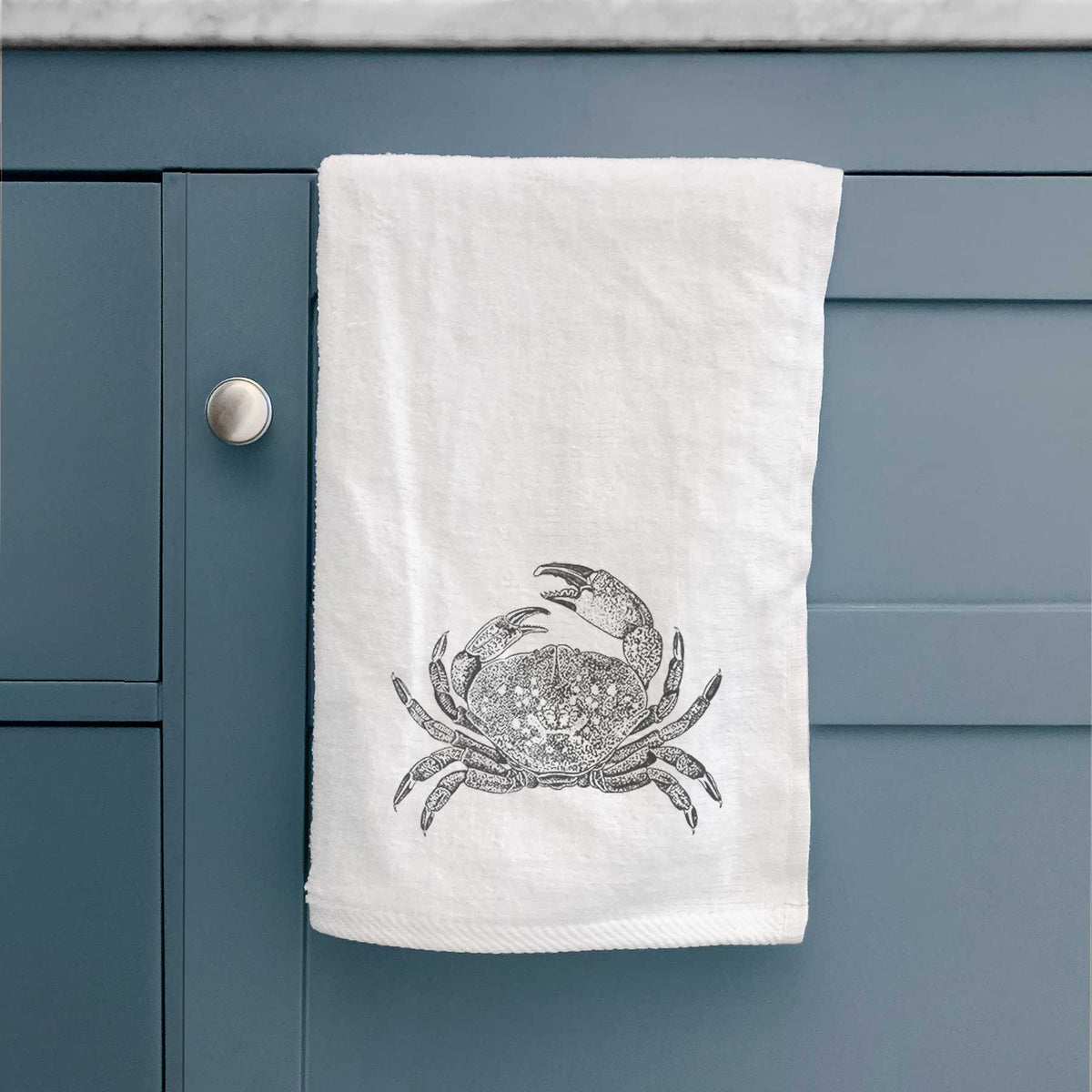 White hand towel printed with a detailed black ink illustration of a Dungeness crab with claws raised, seen from above, hanging on a cabinet door.