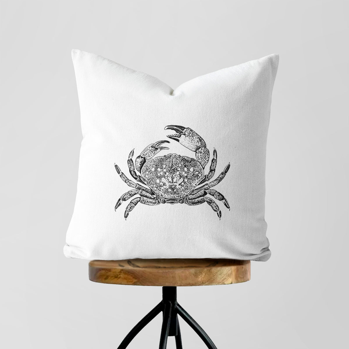 White pillow cover printed with a detailed black ink illustration of a Dungeness crab with claws raised, seen from above, on a wooden stool.