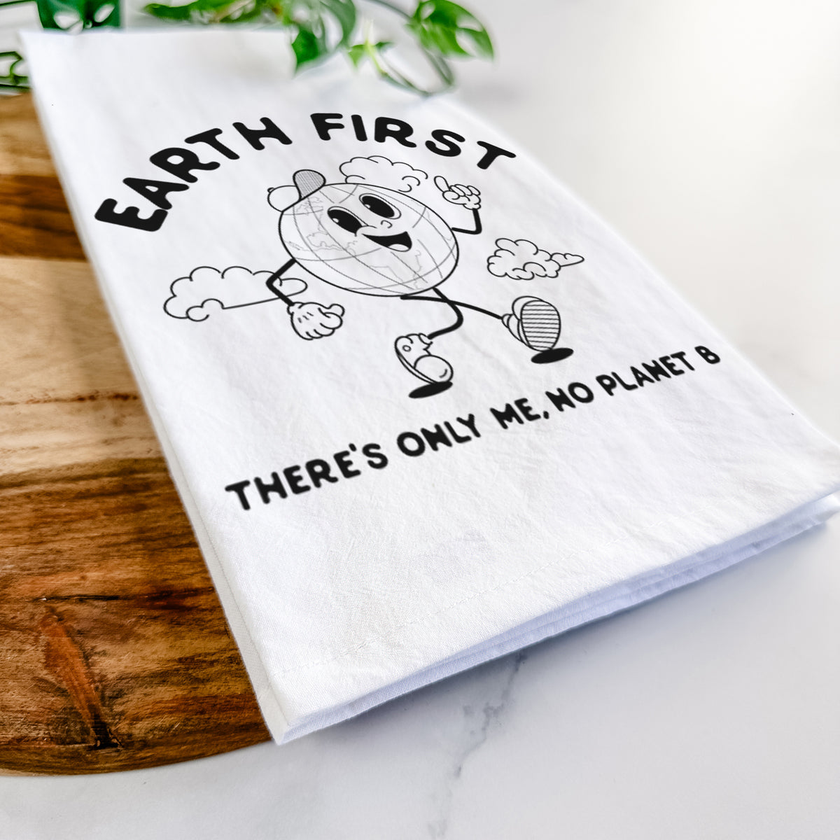 Close-up of a folded white tea towel on a wooden board, showing a cheerful cartoon Earth running, with the words Earth First and There's Only Me, No Planet B.
