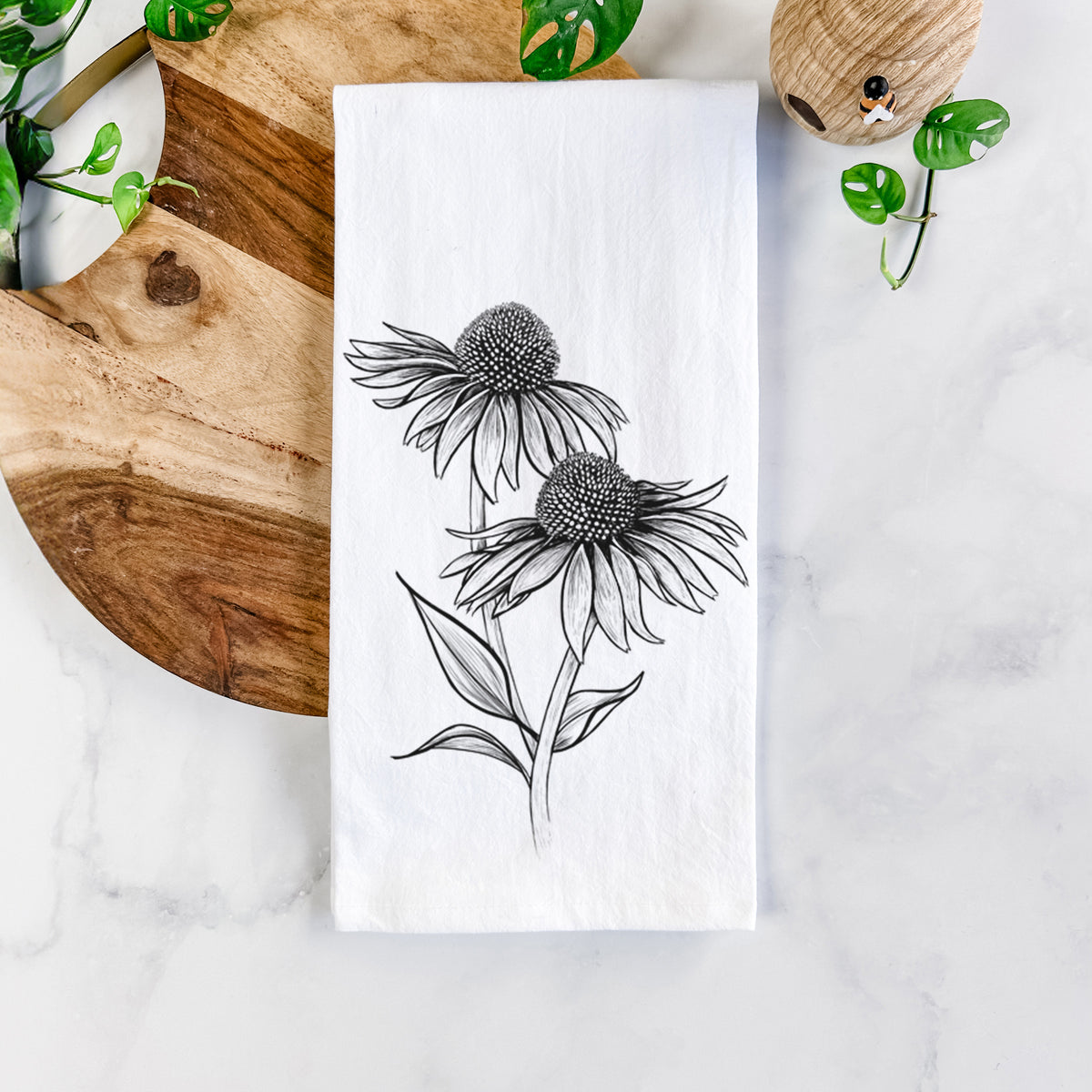 White cotton tea towel printed with a black ink line drawing of two coneflowers on tall stems, on a marble counter beside a round wooden cutting board and greenery.