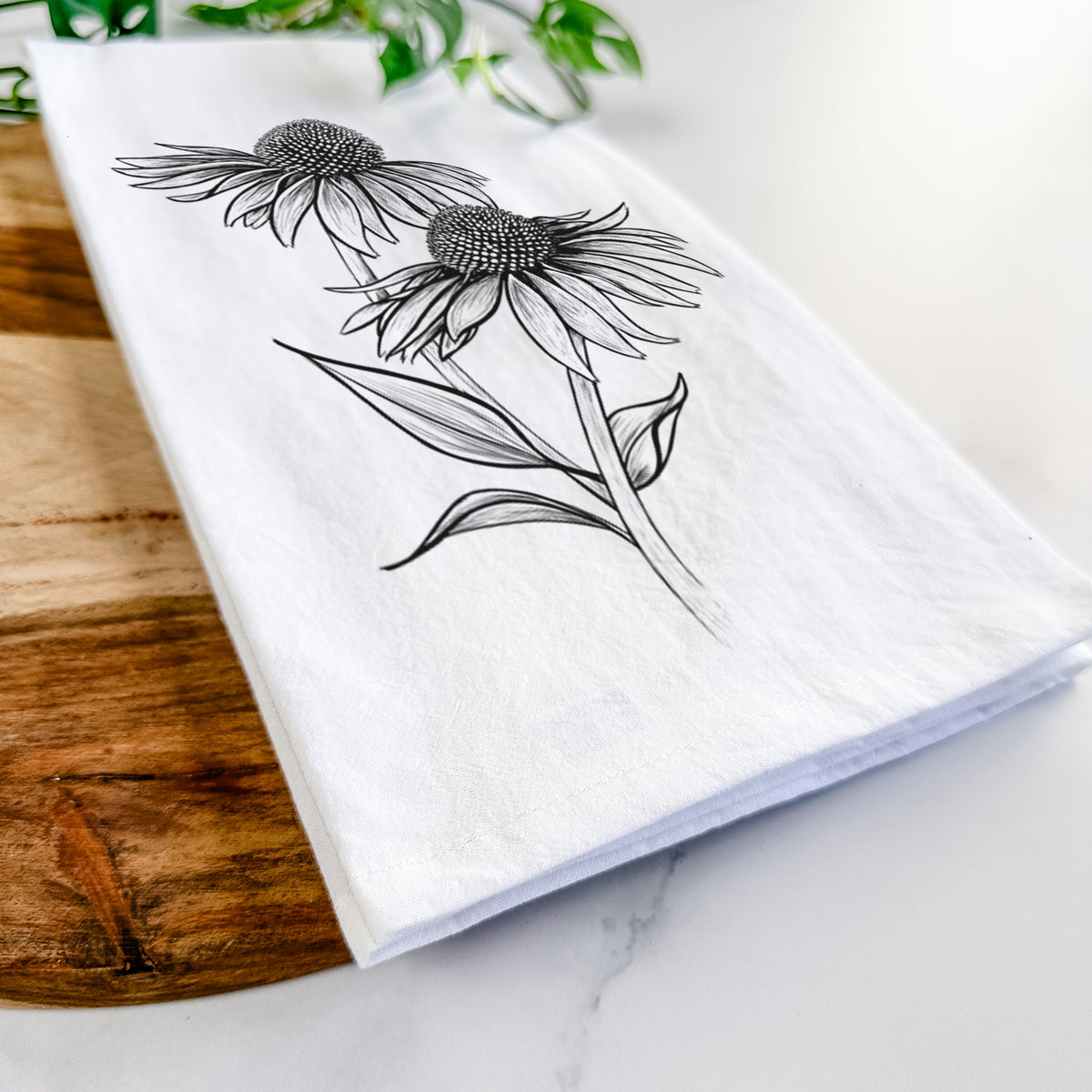 Close-up of a folded white tea towel on a wooden board, showing a black ink line drawing of two coneflowers on tall stems.