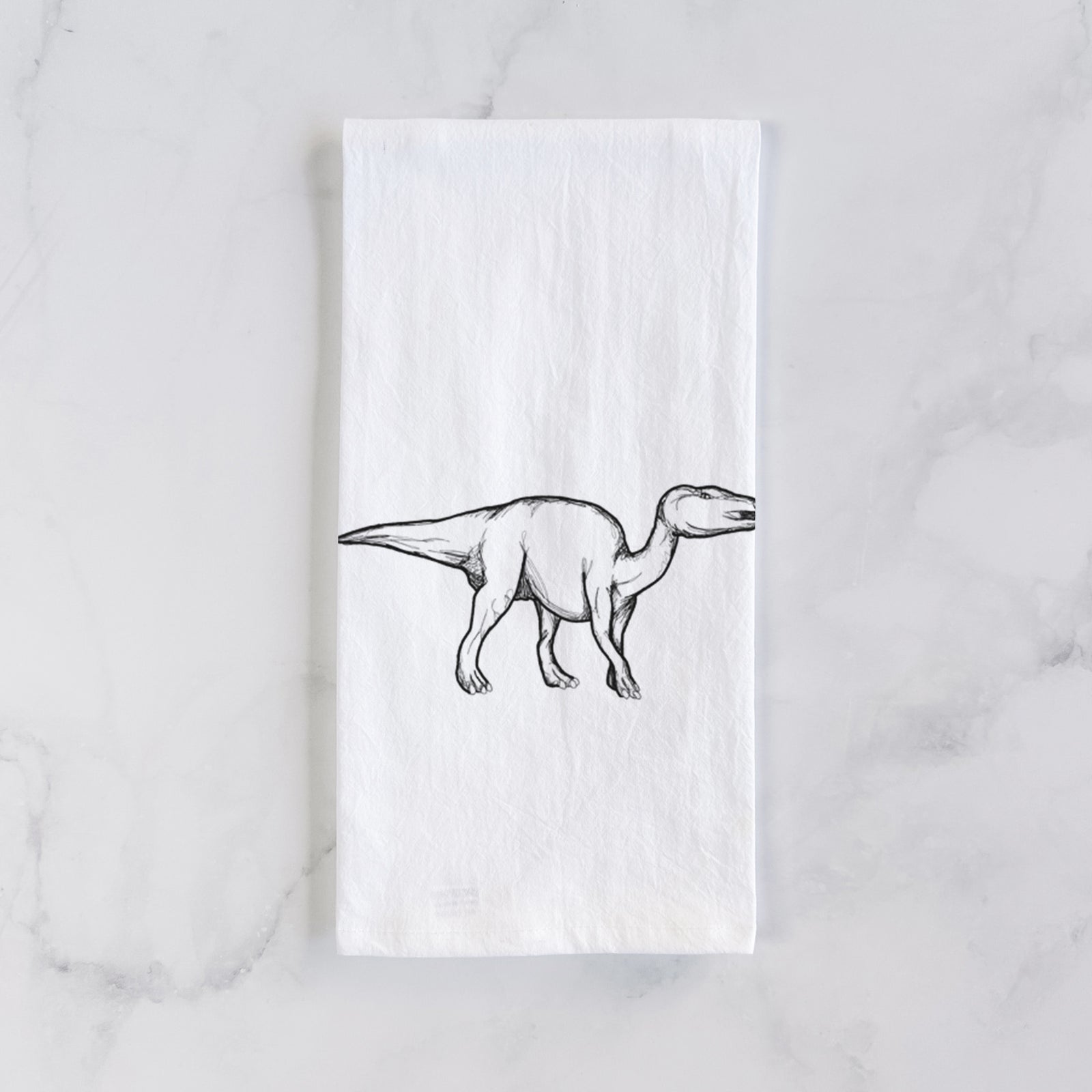 White cotton tea towel printed with a black ink illustration of an Edmontosaurus walking, on a marble counter beside a round wooden cutting board and greenery.