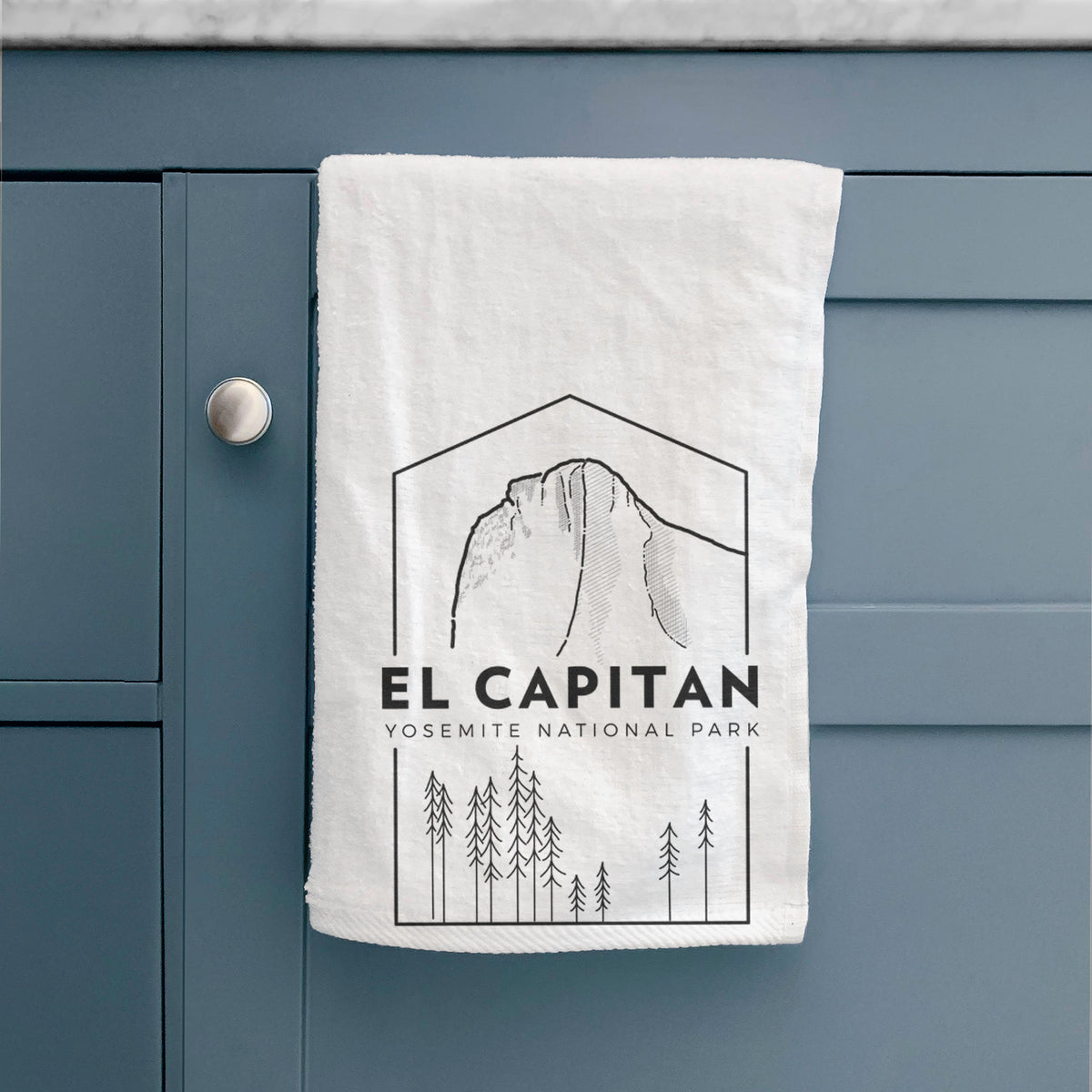 White hand towel printed with a black line art badge of El Capitan rising above a pine forest, with the words El Capitan, Yosemite National Park, hanging on a cabinet door.