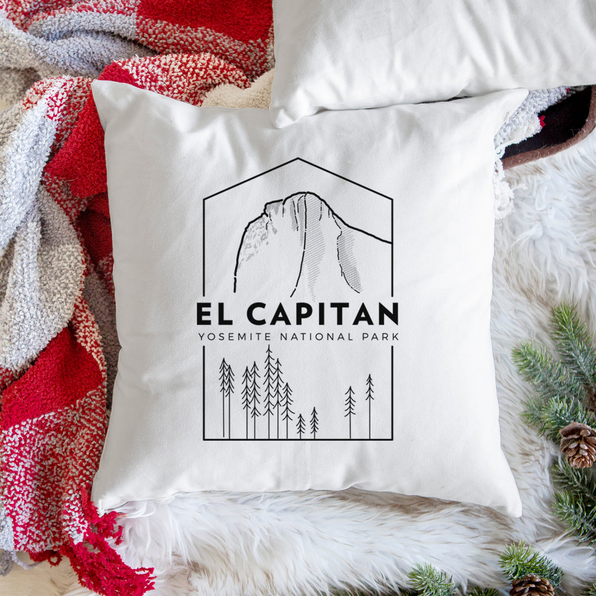 White pillow cover printed with a black line art badge of El Capitan rising above a pine forest, with the words El Capitan, Yosemite National Park, on a woven chair.