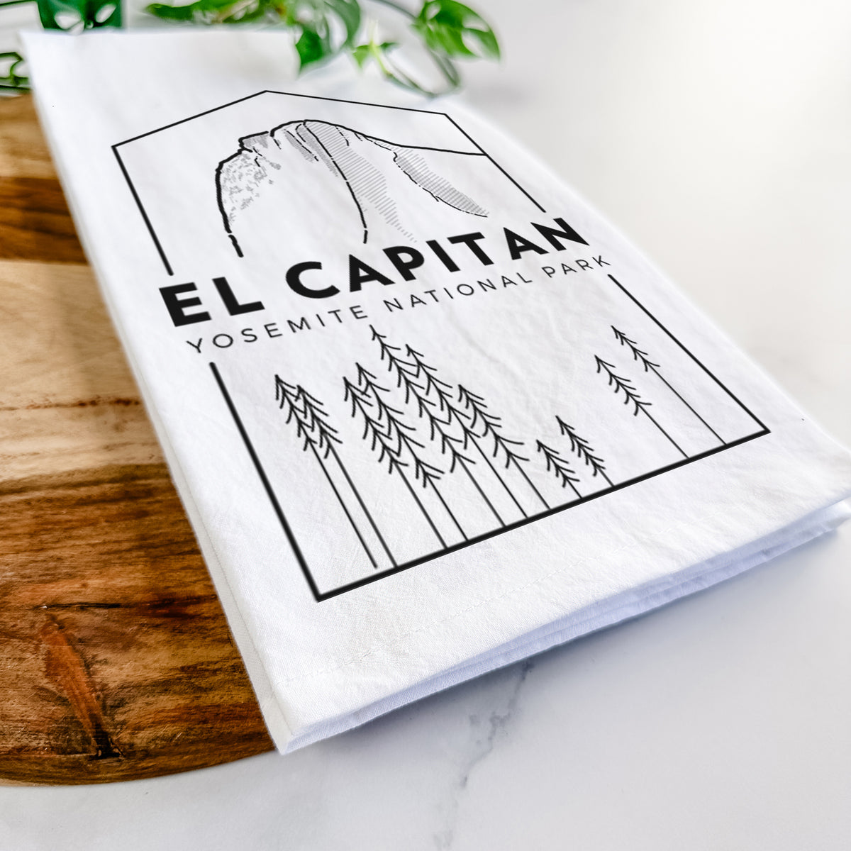Close-up of a folded white tea towel on a wooden board, showing a black line art badge of El Capitan rising above a pine forest, with the words El Capitan, Yosemite National Park.