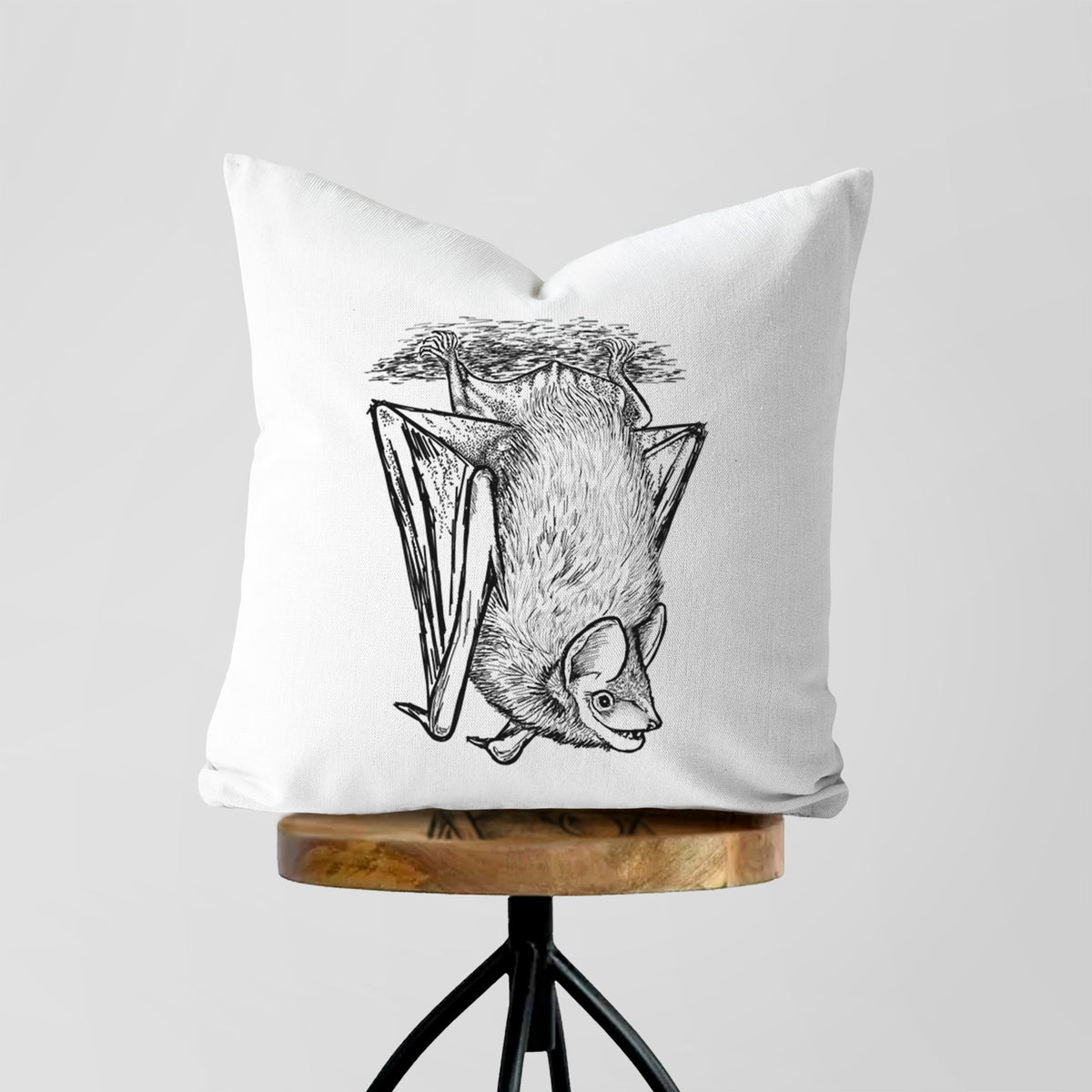 White pillow cover printed with a detailed black ink illustration of a big brown bat hanging upside down, on a wooden stool.