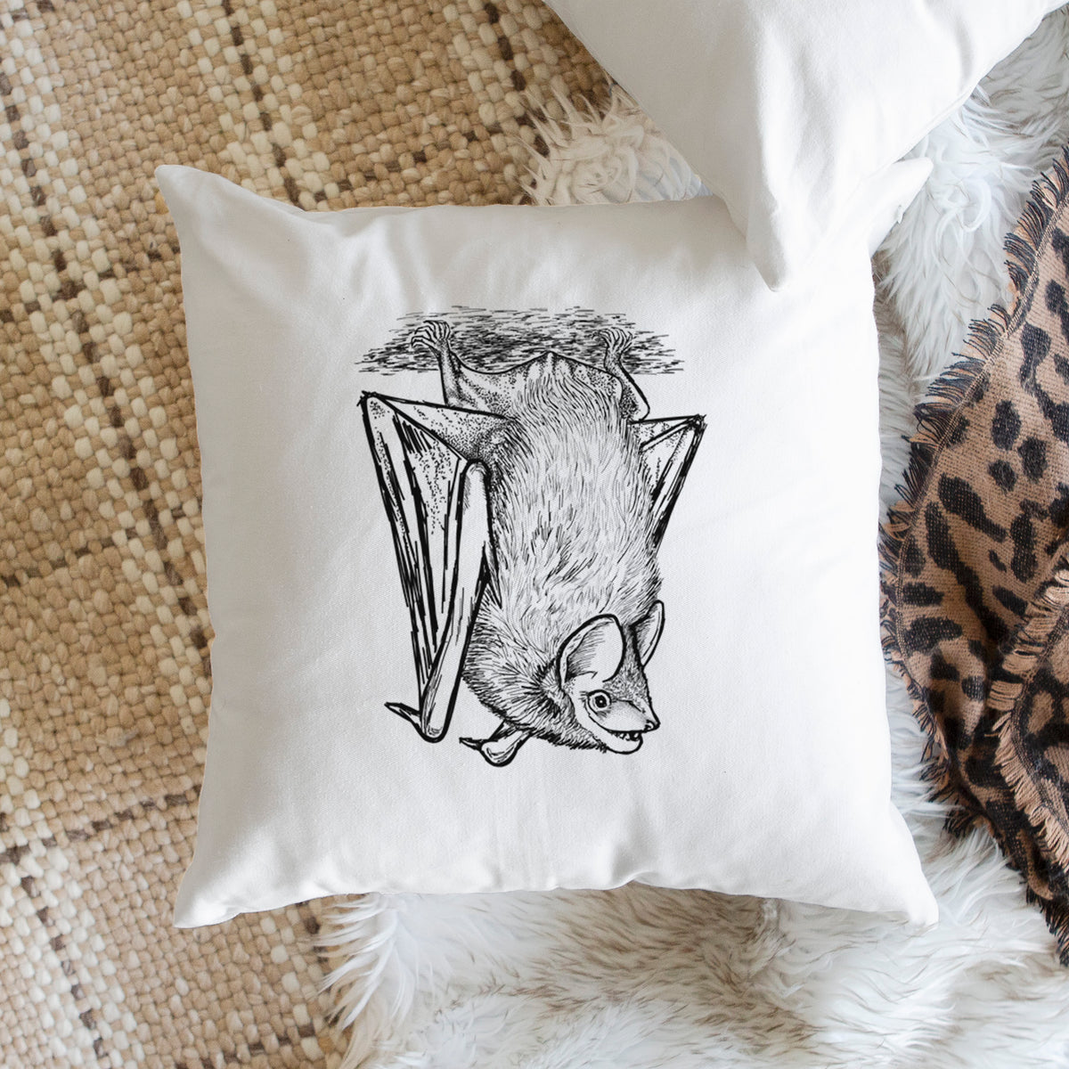 White pillow cover printed with a detailed black ink illustration of a big brown bat hanging upside down, on a woven chair.