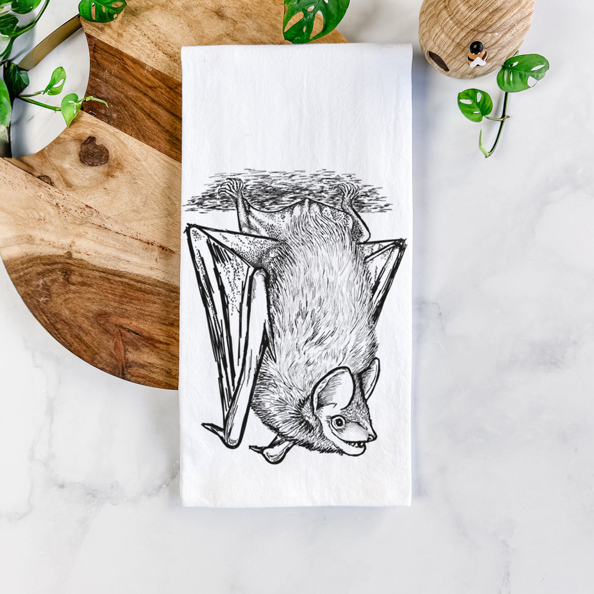 White cotton tea towel printed with a detailed black ink illustration of a big brown bat hanging upside down, on a marble counter beside a round wooden cutting board and greenery.