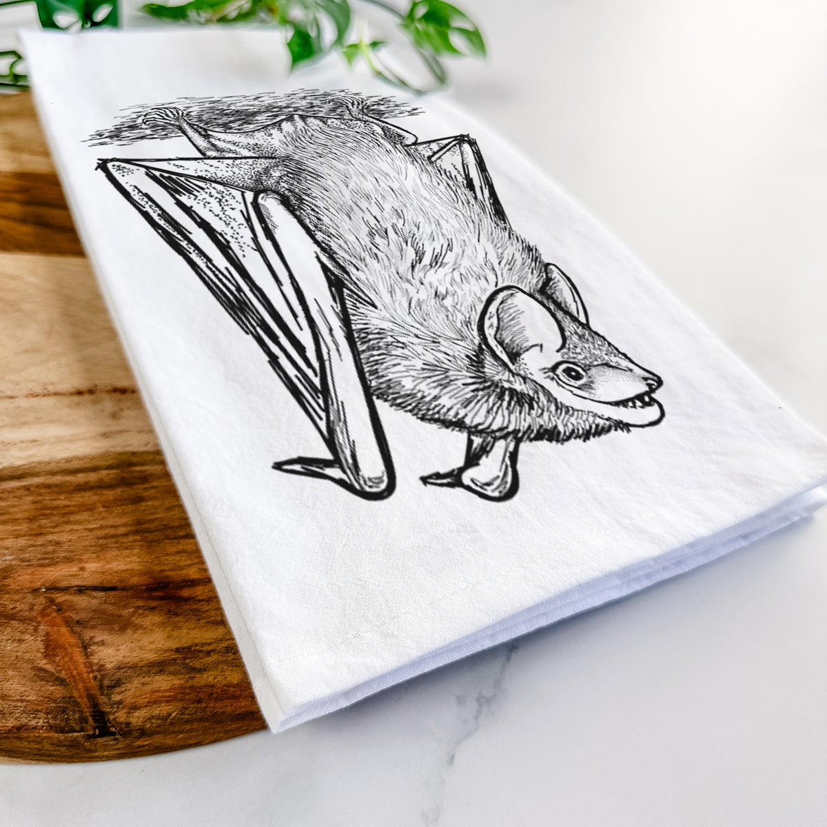 Close-up of a folded white tea towel on a wooden board, showing a detailed black ink illustration of a big brown bat hanging upside down.