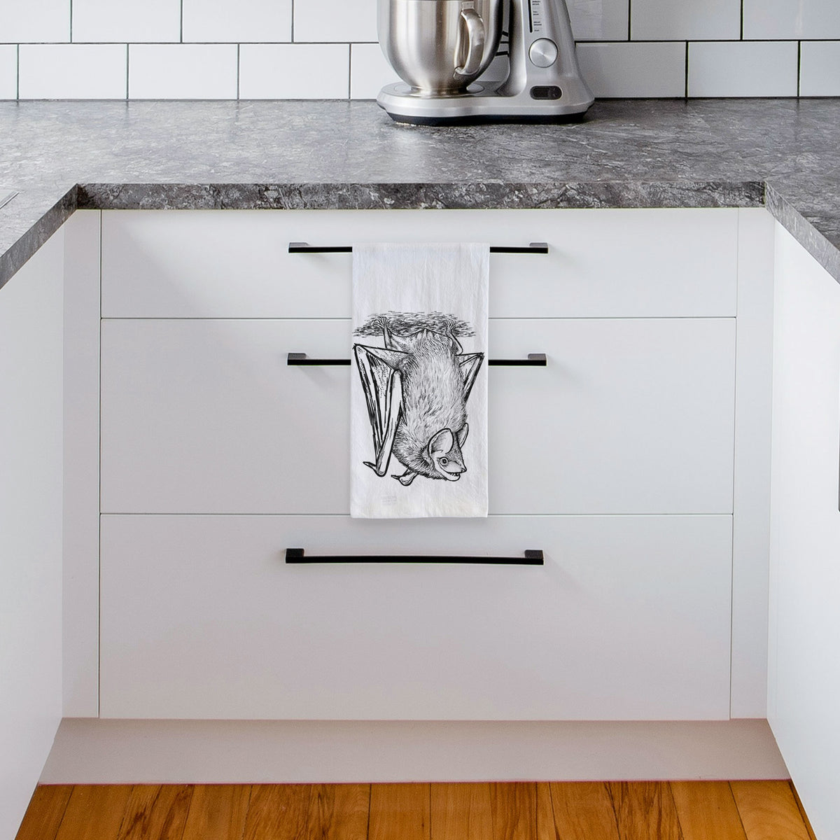 White tea towel printed with a detailed black ink illustration of a big brown bat hanging upside down, hanging from a drawer handle in a bright white kitchen.