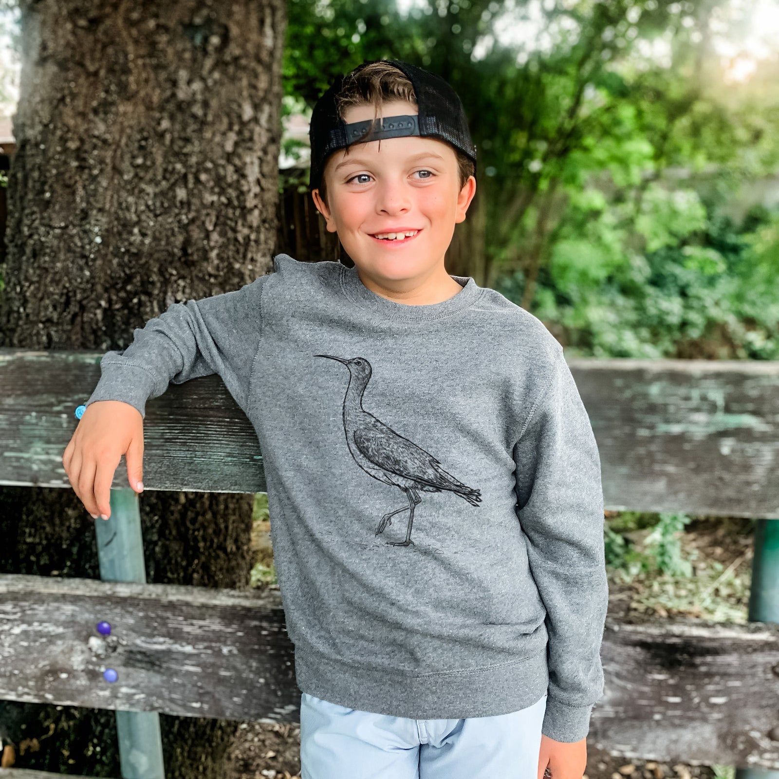 Boy wearing a youth crewneck sweatshirt printed with a detailed black ink illustration of an Eskimo curlew with its long curved bill, walking, outdoors.