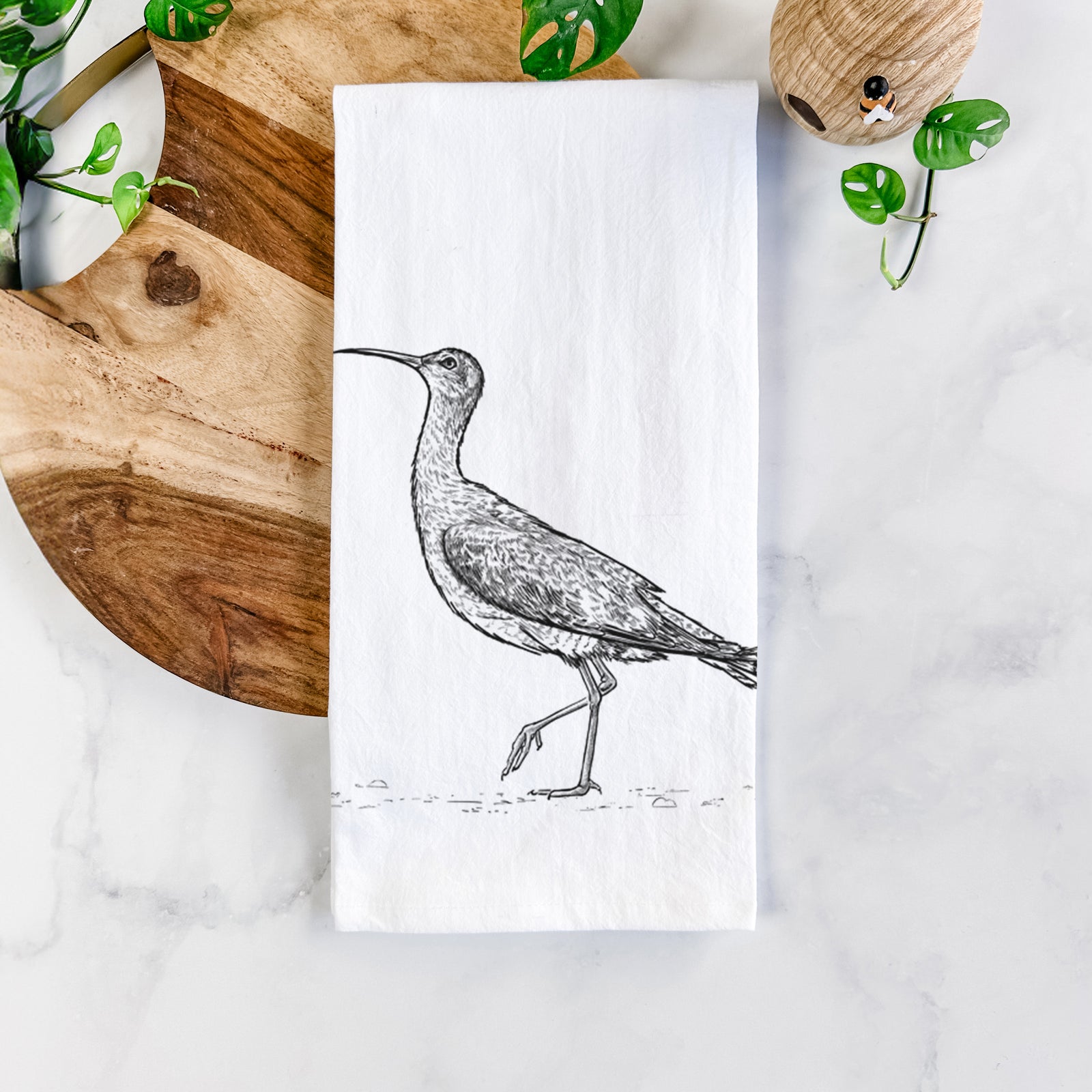 White cotton tea towel printed with a detailed black ink illustration of an Eskimo curlew with its long curved bill, walking, on a marble counter beside a round wooden cutting board and greenery.