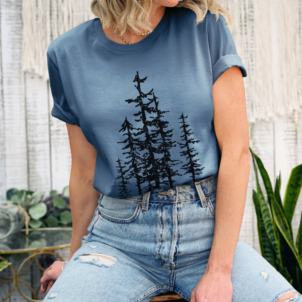 Woman wearing a steel blue lightweight cotton t-shirt printed with a black ink illustration of a cluster of evergreen trees of varying heights.