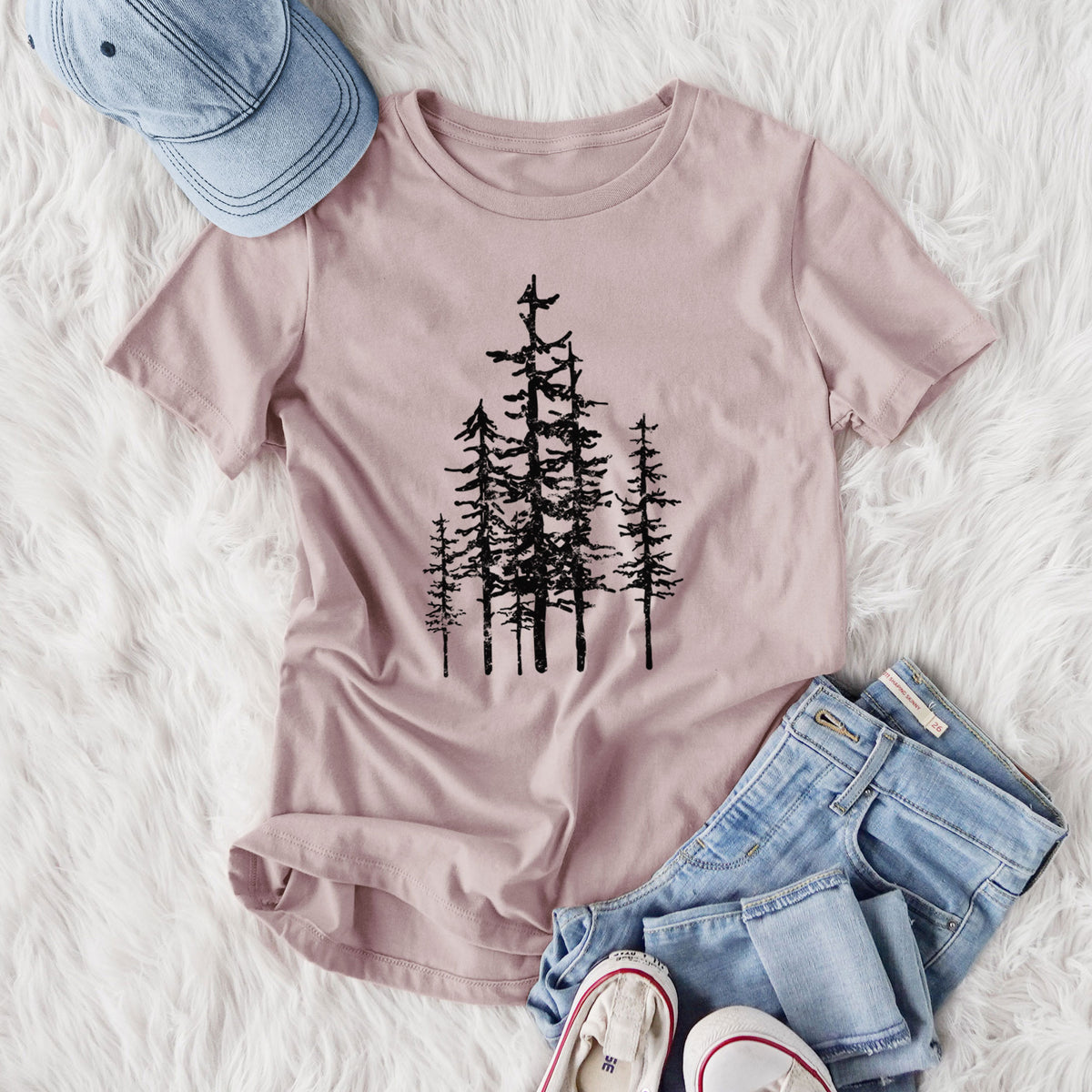 Women's relaxed fit cotton t-shirt printed with a black ink illustration of a cluster of evergreen trees of varying heights, styled flat on a white faux fur rug with jeans.
