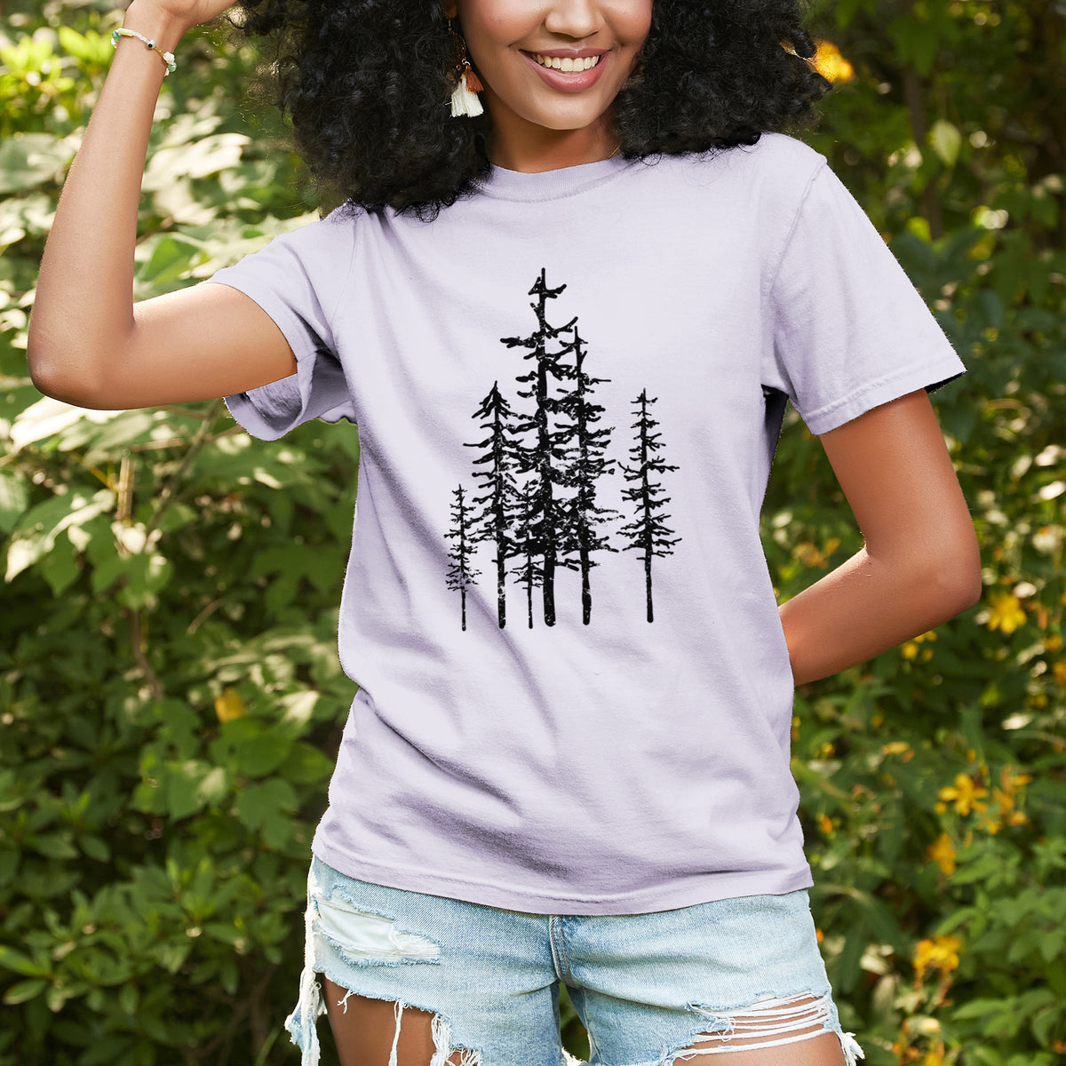 Woman wearing a Comfort Colors t-shirt printed with a black ink illustration of a cluster of evergreen trees of varying heights, outdoors.