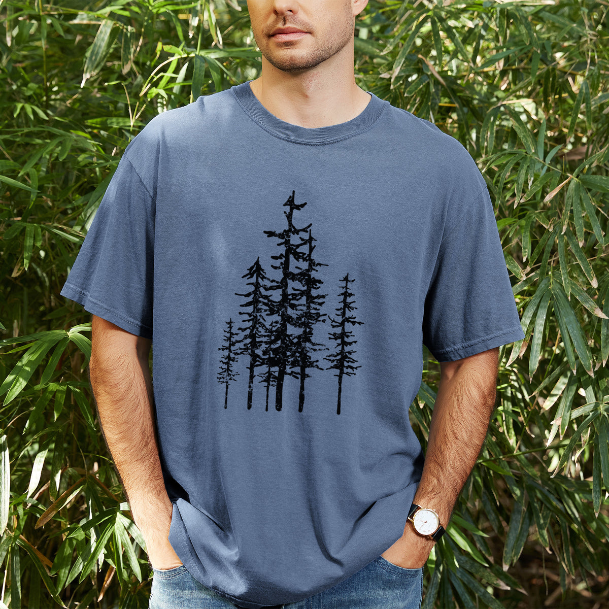 Man wearing a Comfort Colors t-shirt printed with a black ink illustration of a cluster of evergreen trees of varying heights, outdoors.