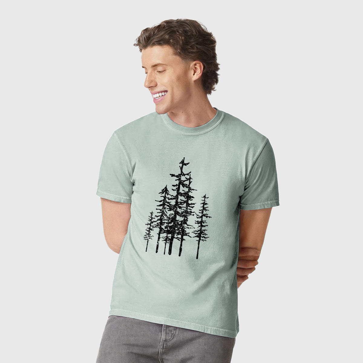 Man wearing a Comfort Colors t-shirt printed with a black ink illustration of a cluster of evergreen trees of varying heights.