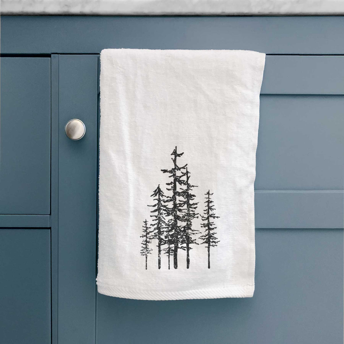 White hand towel printed with a black ink illustration of a cluster of evergreen trees of varying heights, hanging on a cabinet door.