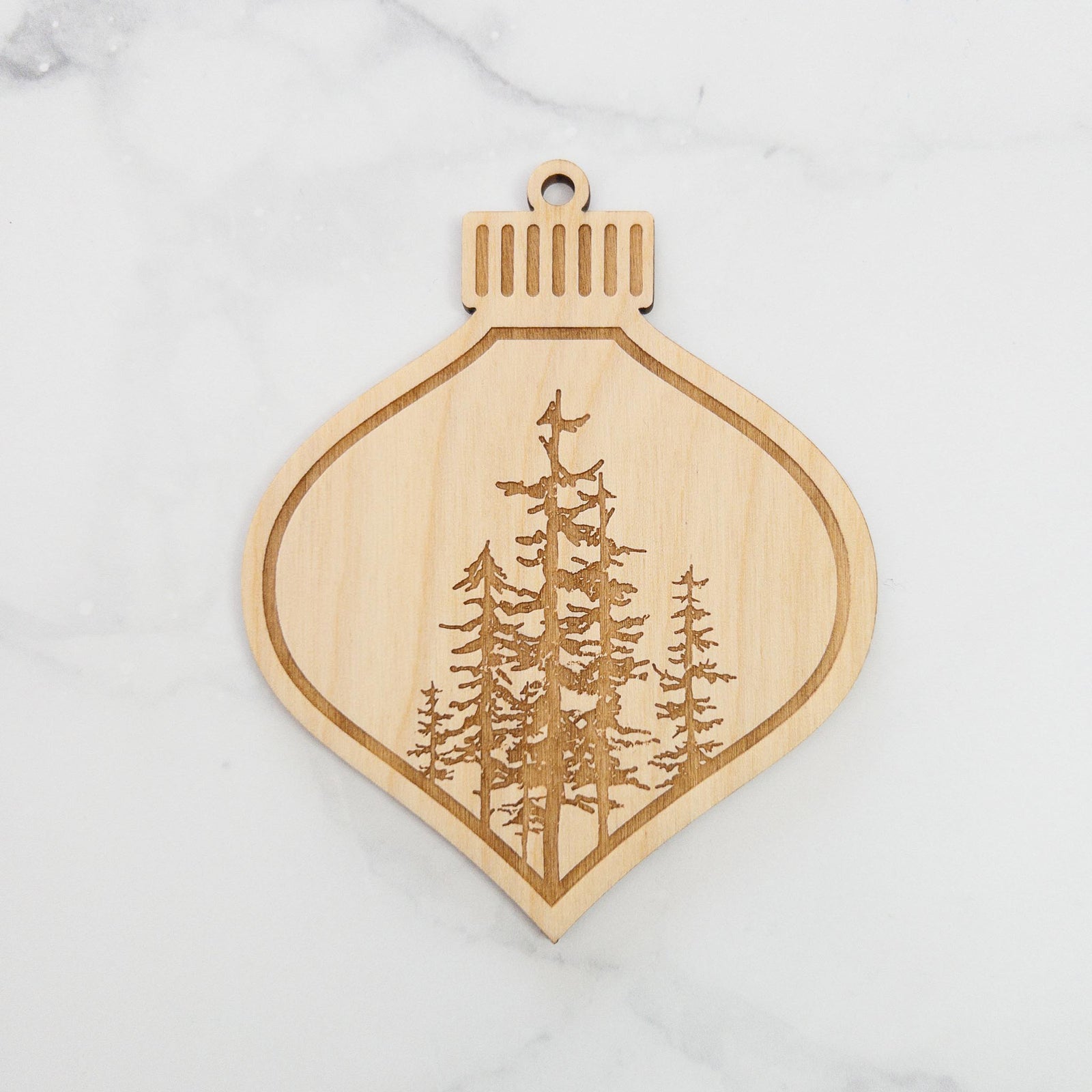 Wooden ornament featuring a engraving of a cluster of evergreen trees of varying heights, on a knit blanket with pine cones.