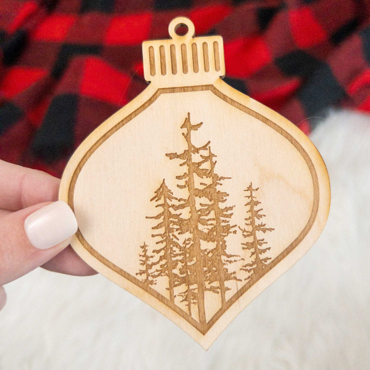 Wooden ornament featuring a engraving of a cluster of evergreen trees of varying heights, on a knit blanket with pine cones.