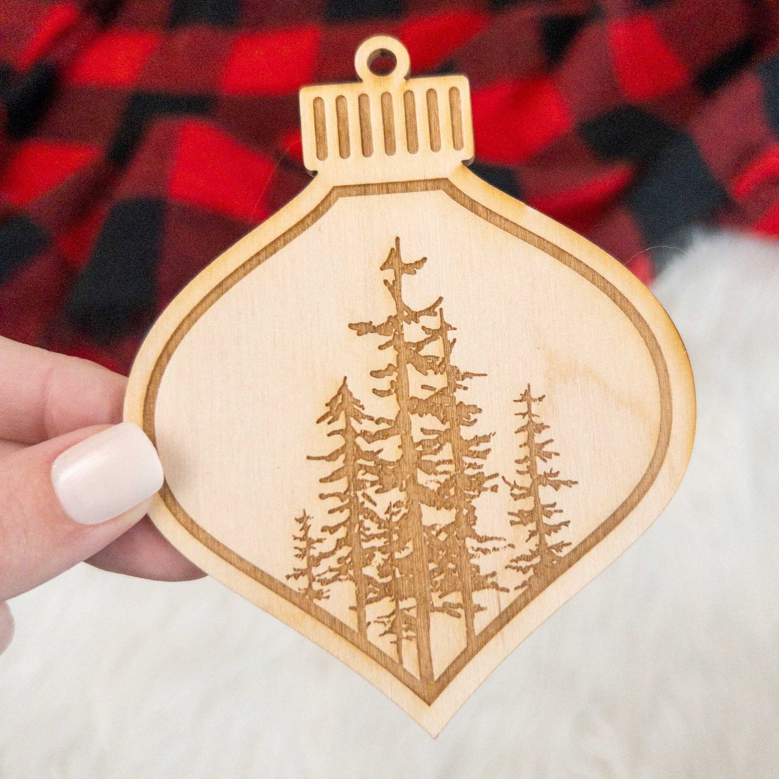Wooden ornament featuring a engraving of a cluster of evergreen trees of varying heights, on a knit blanket with pine cones.