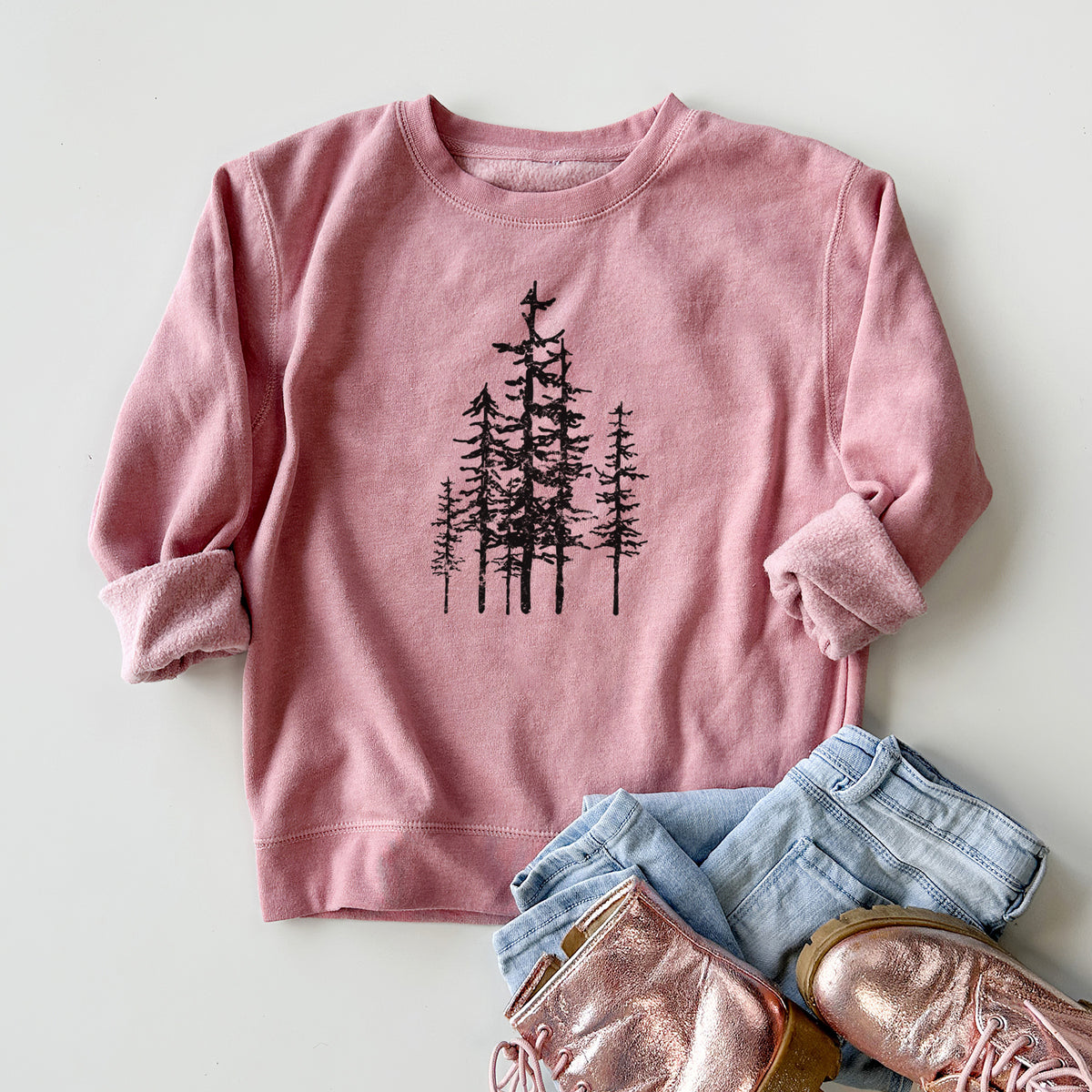 Youth crewneck sweatshirt printed with a black ink illustration of a cluster of evergreen trees of varying heights, styled flat with jeans and boots.