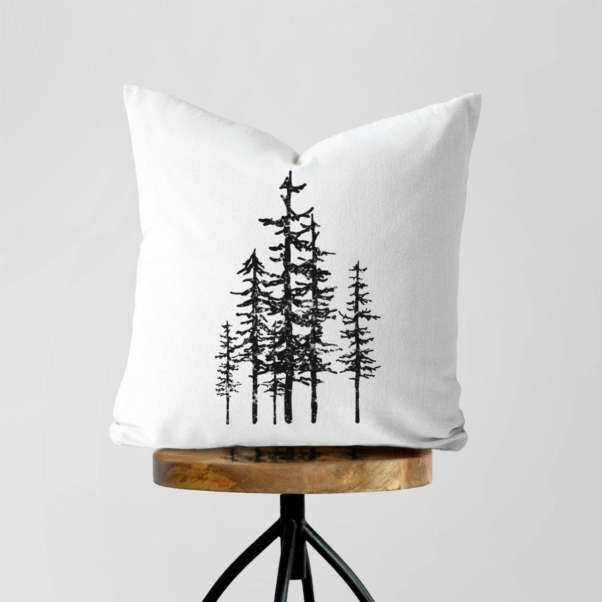 White pillow cover printed with a black ink illustration of a cluster of evergreen trees of varying heights, on a wooden stool.